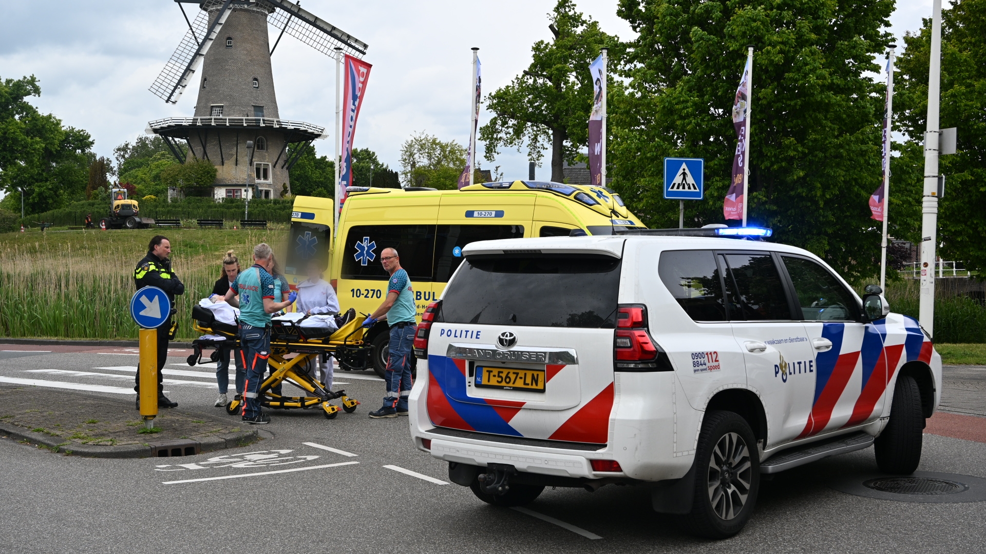 Een ambulance en politieauto staan op een kruispunt bij een historische windmolen, terwijl paramedici een persoon op een brancard behandelen.
