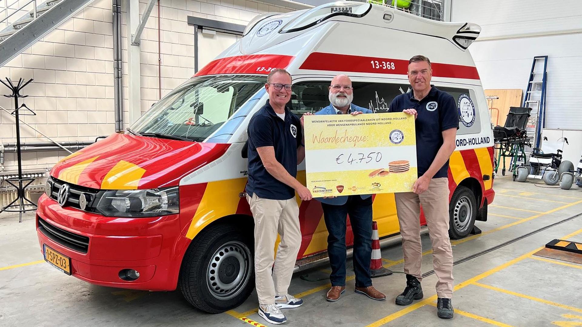 Drie mannen poseren met een cheque van €4.750 voor een wensambulance in rood-geel-blauwe kleuren in een loods.