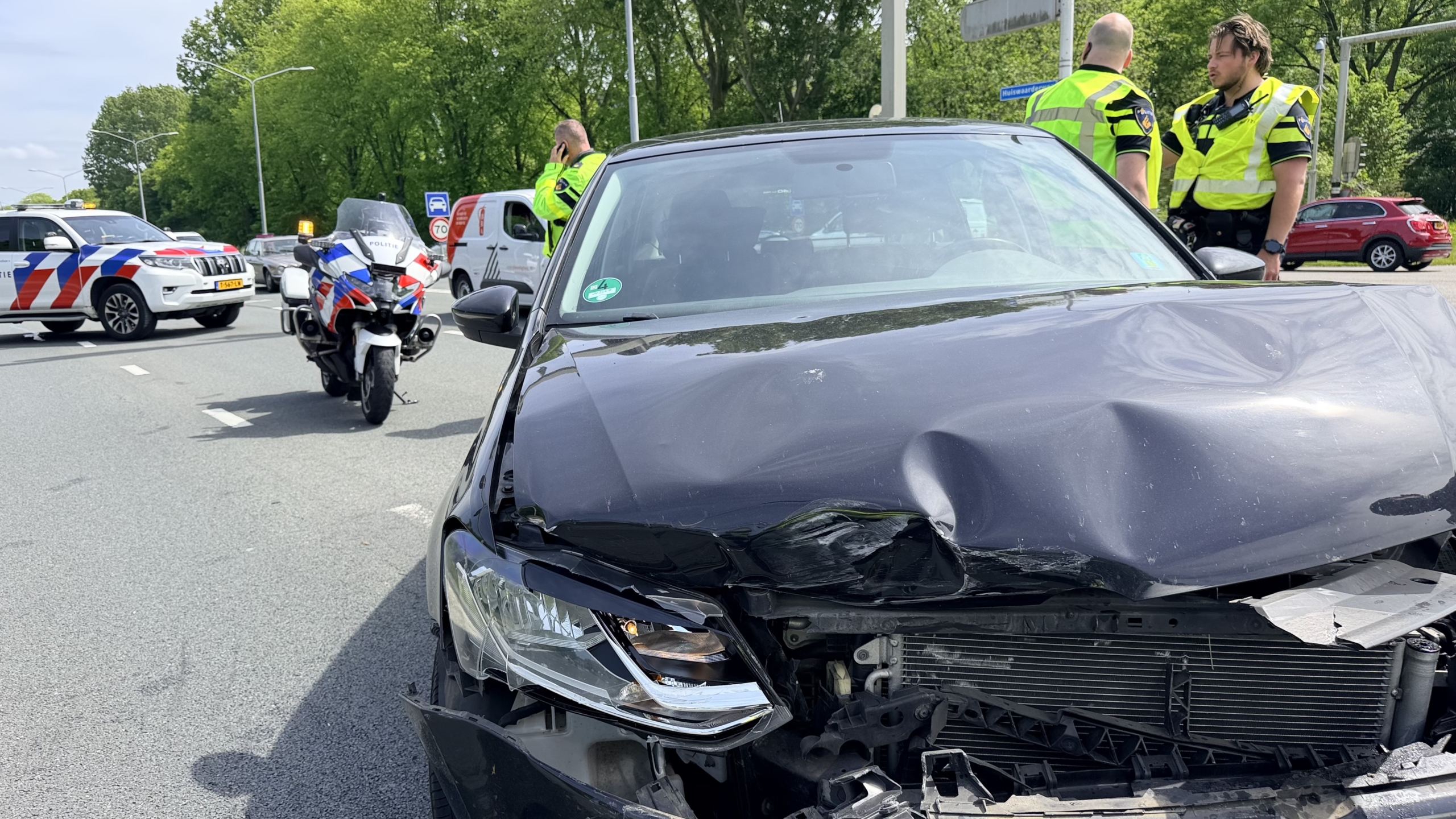Een zwart voertuig met schade aan de voorkant, omringd door politieauto’s en -motorfiets, met agenten in gesprek op de achtergrond.