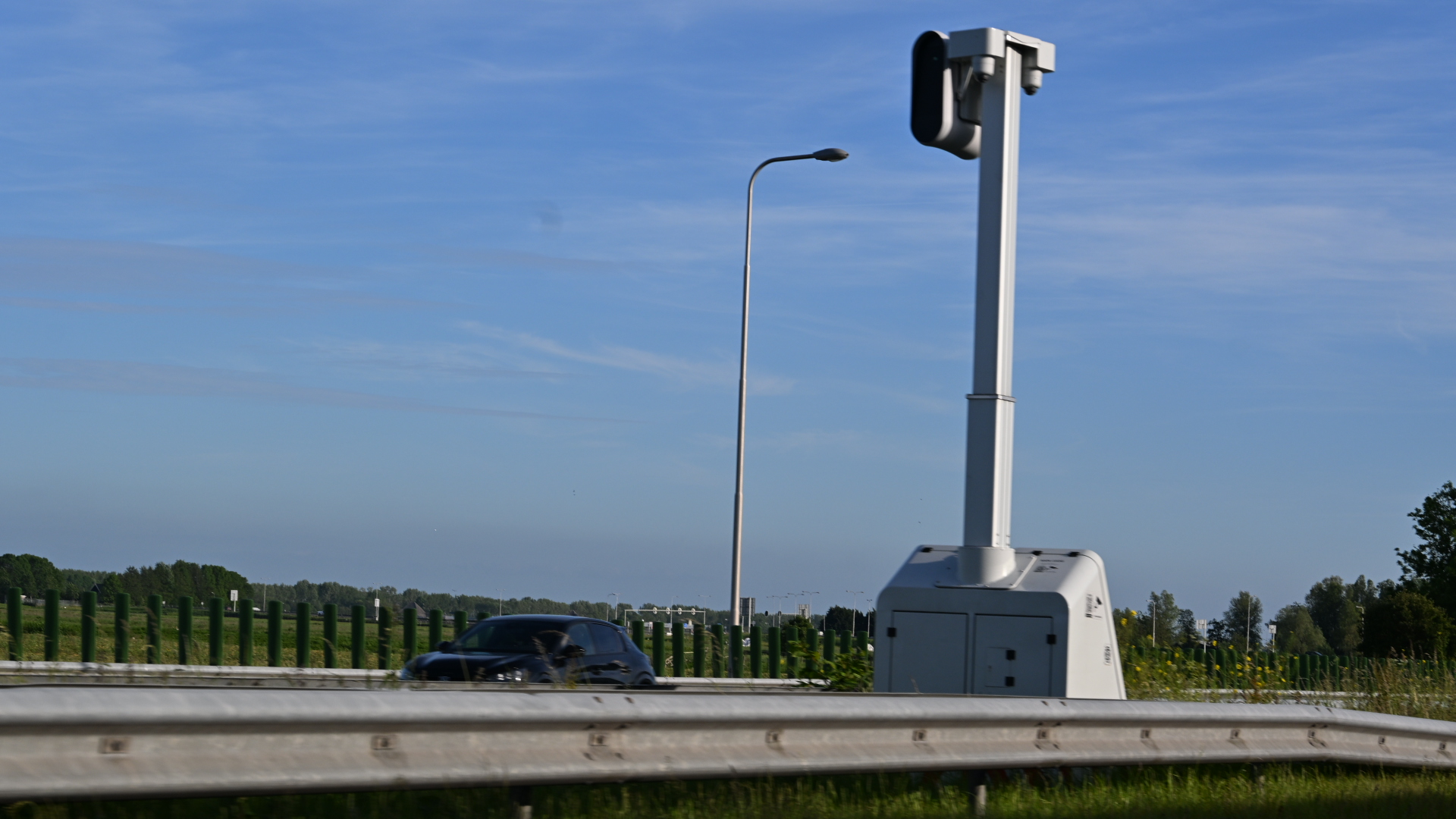 Verkeerscamera langs een snelweg met een auto die voorbijrijdt onder een heldere blauwe hemel.