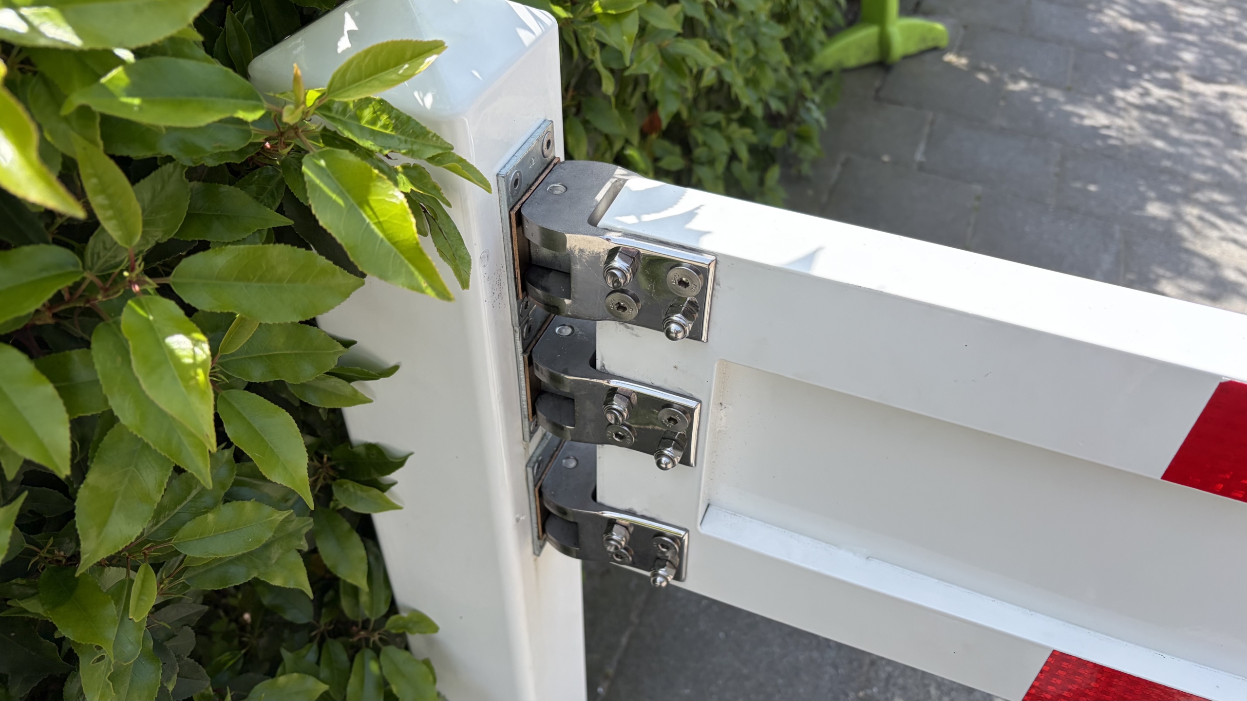 Close-up van een metalen scharnier op een wit hek, omringd door groene bladeren en een stoep op de achtergrond.