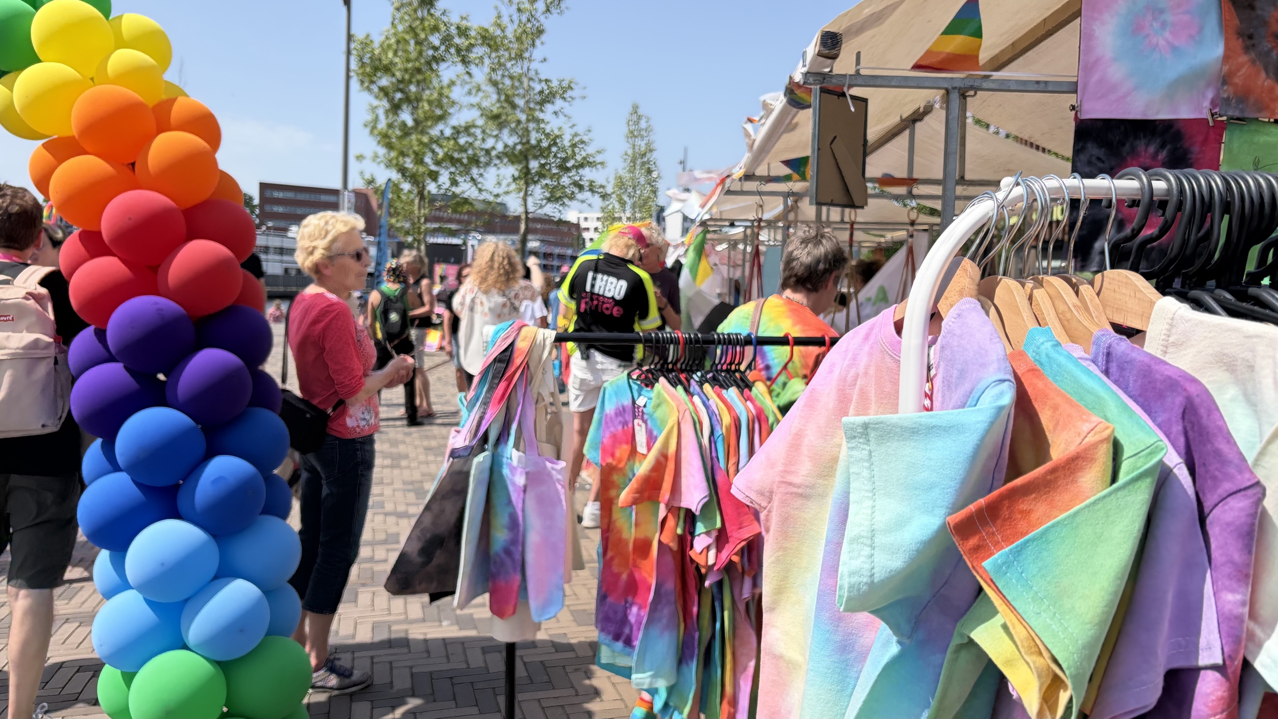 Regenboogballonnen en kleurrijke tie-dye kleding bij een openluchtmarkt op een zonnige dag.