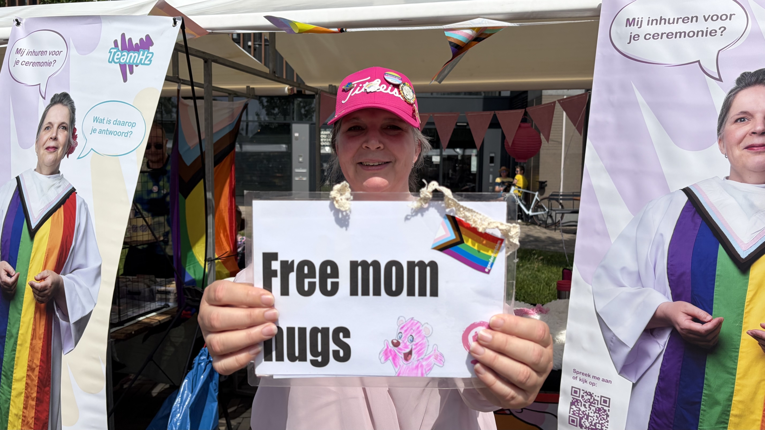 Vrouw met roze pet houdt een bord vast met "Free mom hugs" voor een kraam met regenboogvlaggen en posters.