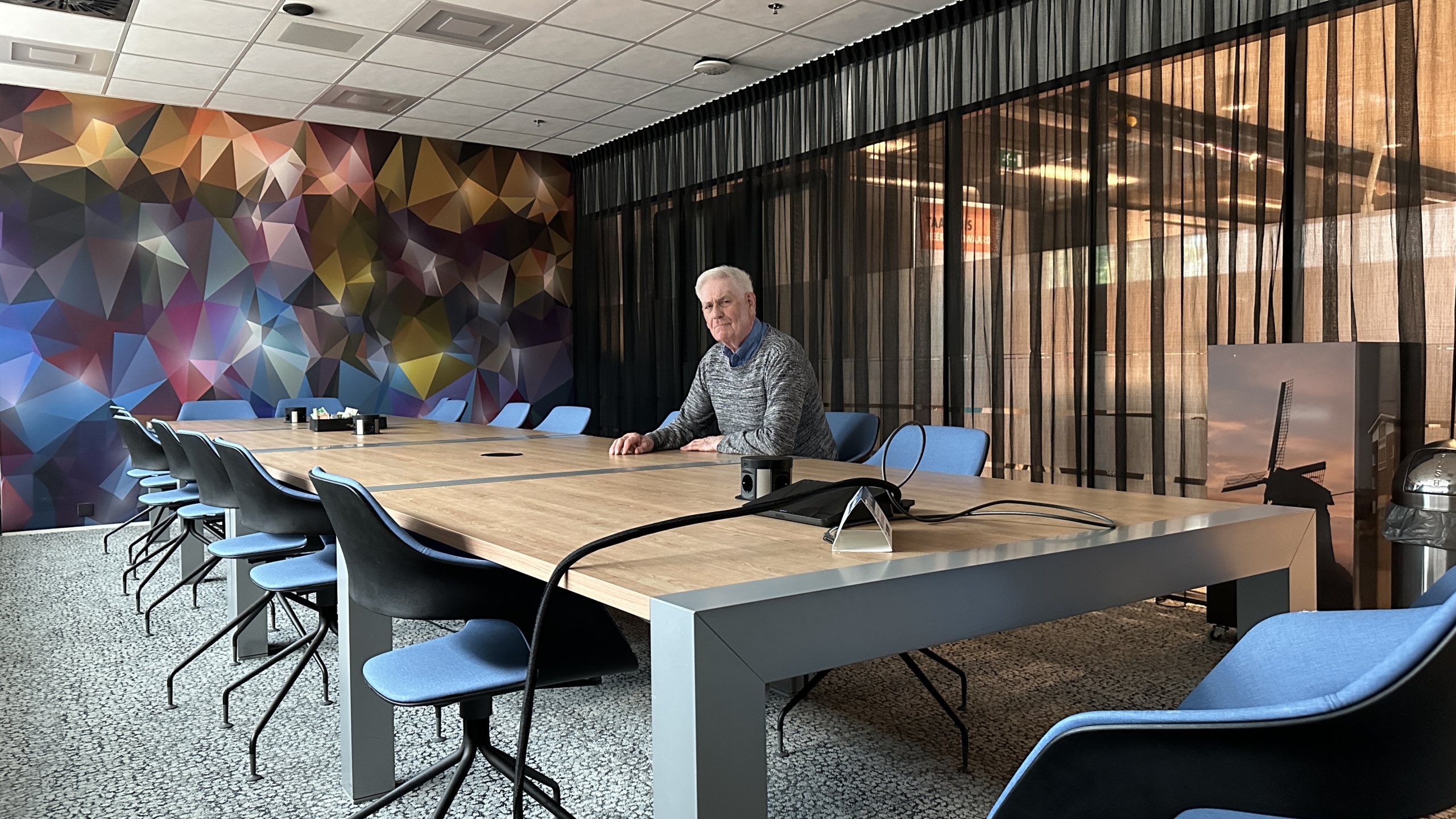 Man zittend aan een vergadertafel in een kleurrijk ingerichte kamer met moderne stoelen en een geometrische muurschildering.