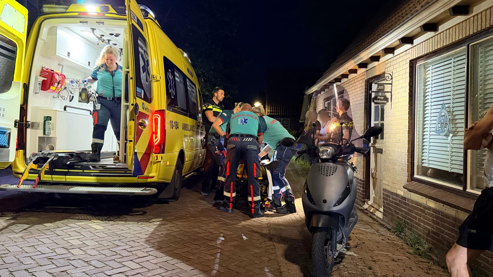 Hulpverleners bij een ambulance verzorgen iemand naast een gebouw, terwijl een scooter op de voorgrond staat.