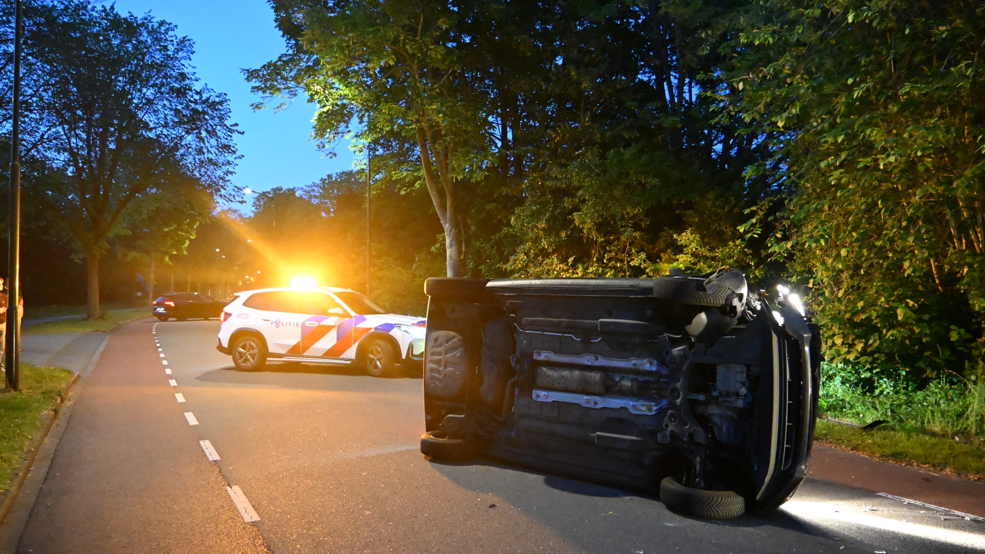 Een gekantelde auto ligt op de weg naast een politieauto met zwaailichten, omgeven door bomen bij schemering.