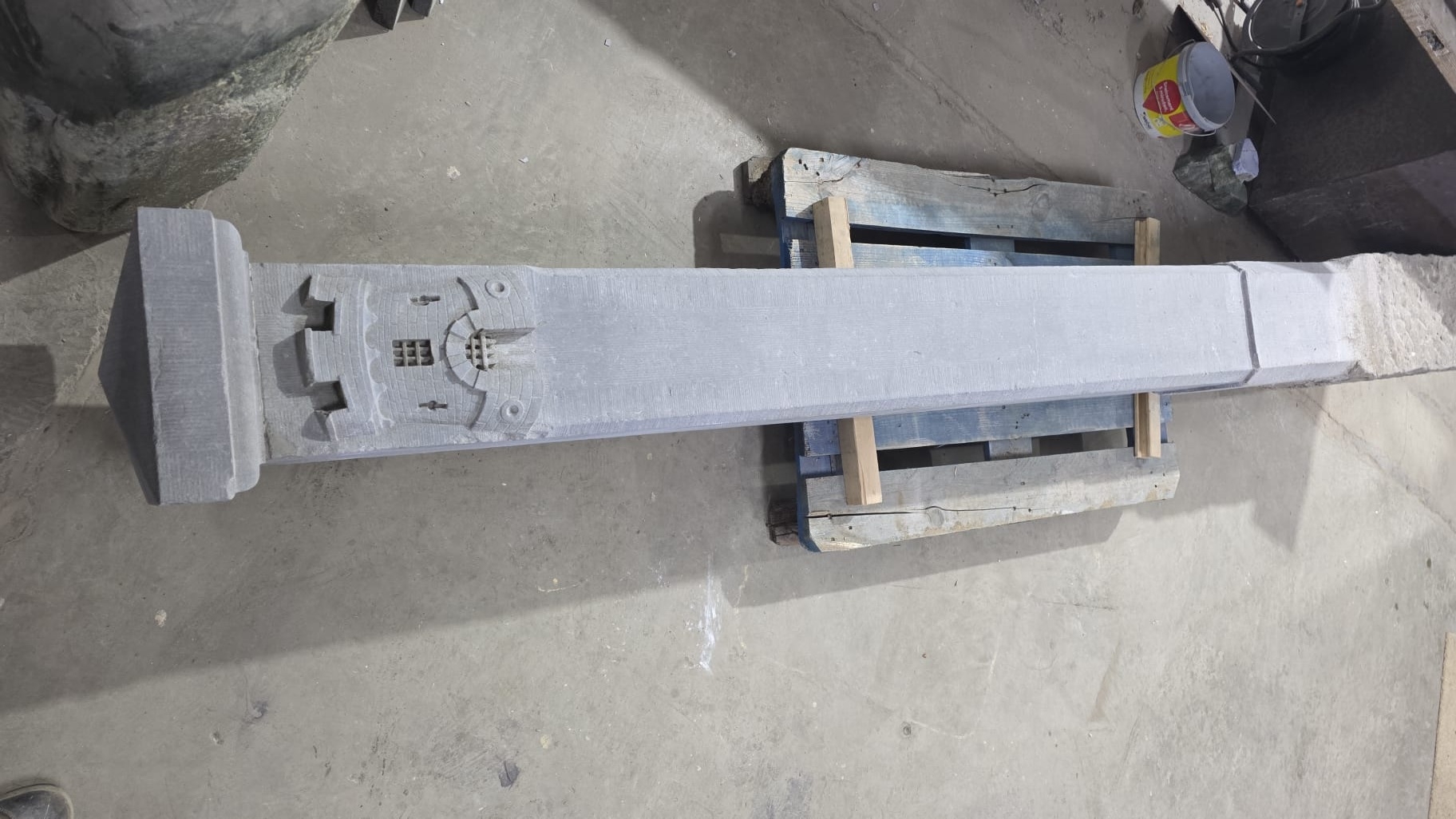 Een lang, rechthoekig stenen object met ingewikkelde gravures, geplaatst op een houten pallet in een werkplaats.