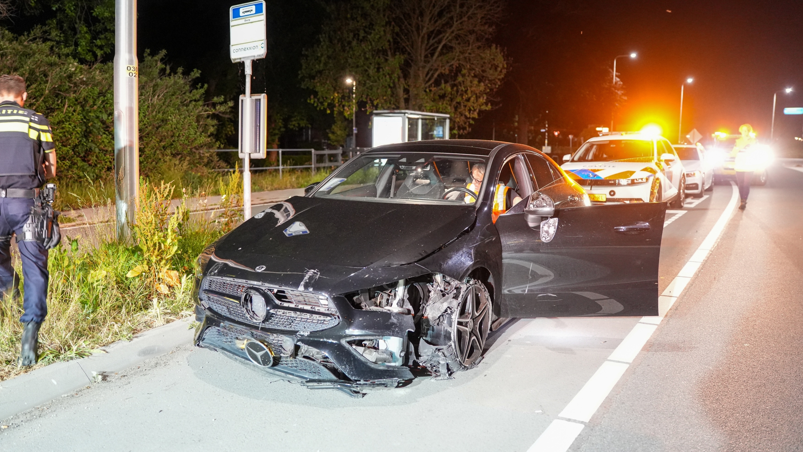 Beschadigde auto langs de weg met geopende deur, politieagent naast de auto en politievoertuig met zwaailicht op de achtergrond.