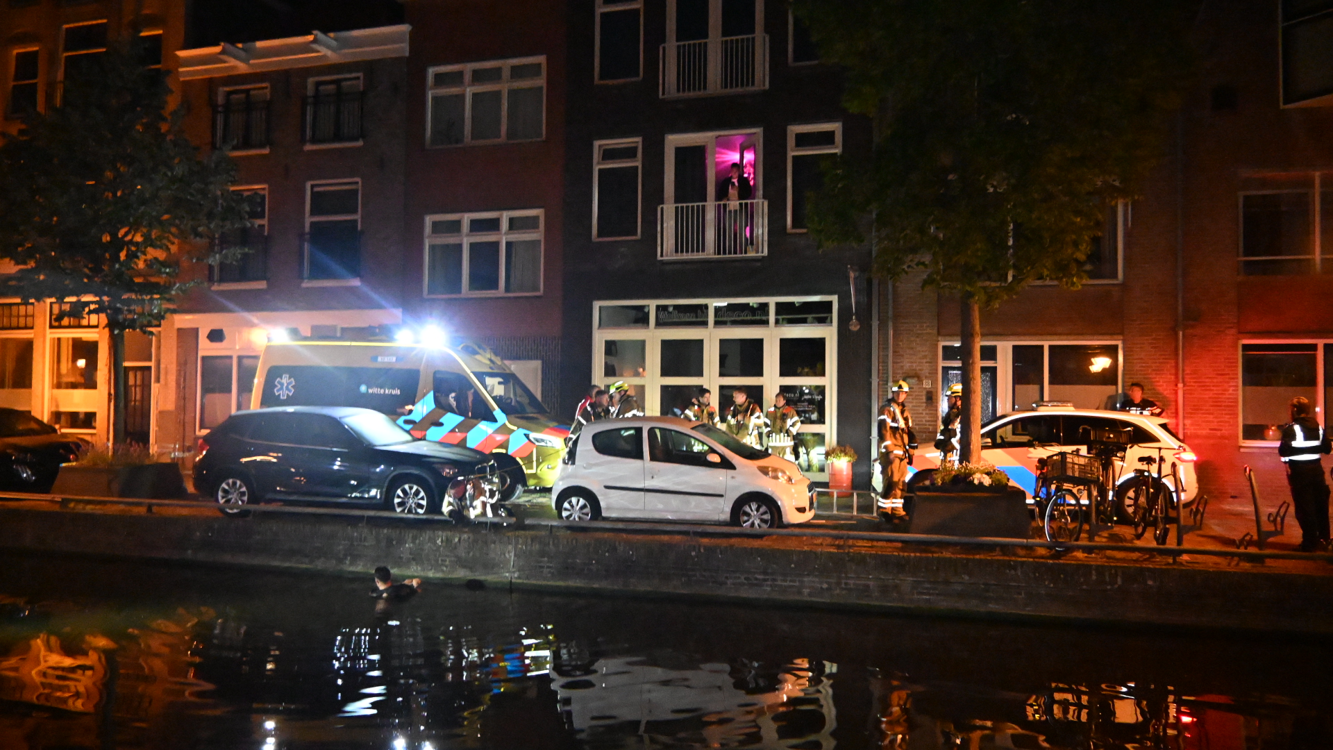 Nachtelijke scène met een ambulance en politieauto voor een gebouw, omringd door hulpverleners, met een persoon in het water.