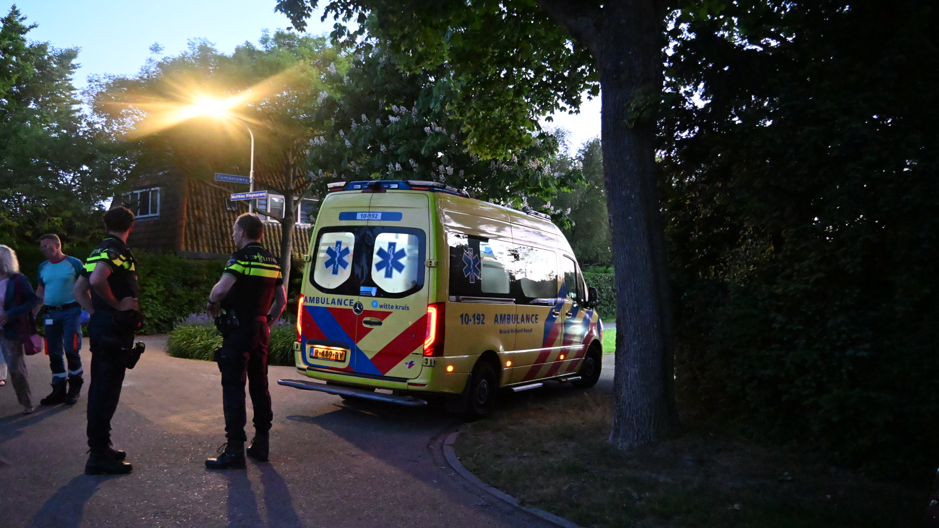 Een ambulance en politieagenten staan geparkeerd naast een boom op een straat verlicht door een lantaarn.