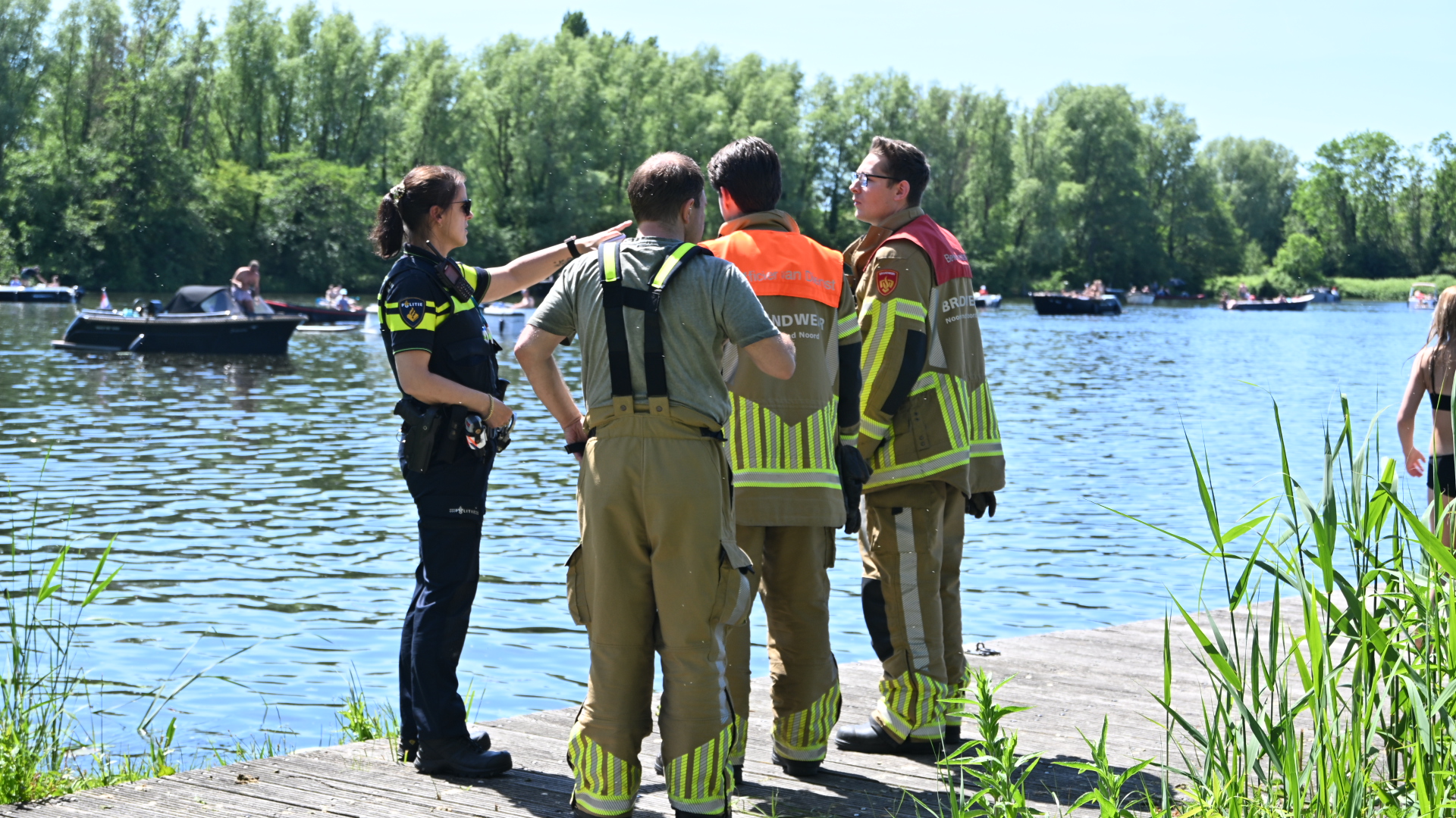 Politie- en brandweermensen staan op een steiger aan een meer, met boten en mensen in de achtergrond.