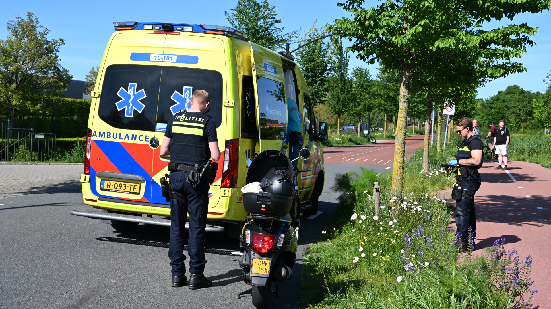 Ambulance en politie bij een incident langs de weg, met een politieagent en een scooter op de voorgrond.