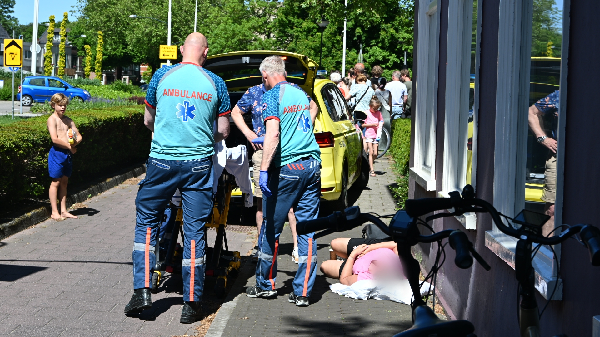 Ambulancepersoneel helpt een persoon op de stoep naast een ambulance; een jongen kijkt toe.