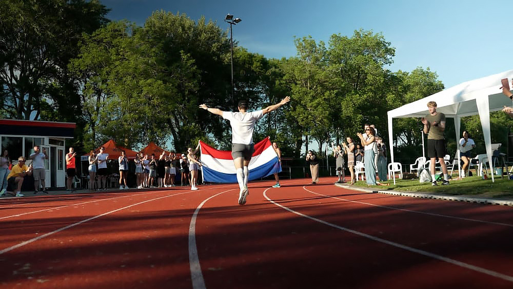 Een hardloper steekt de finishlijn over met gespreide armen, omringd door juichende toeschouwers en een Nederlandse vlag.