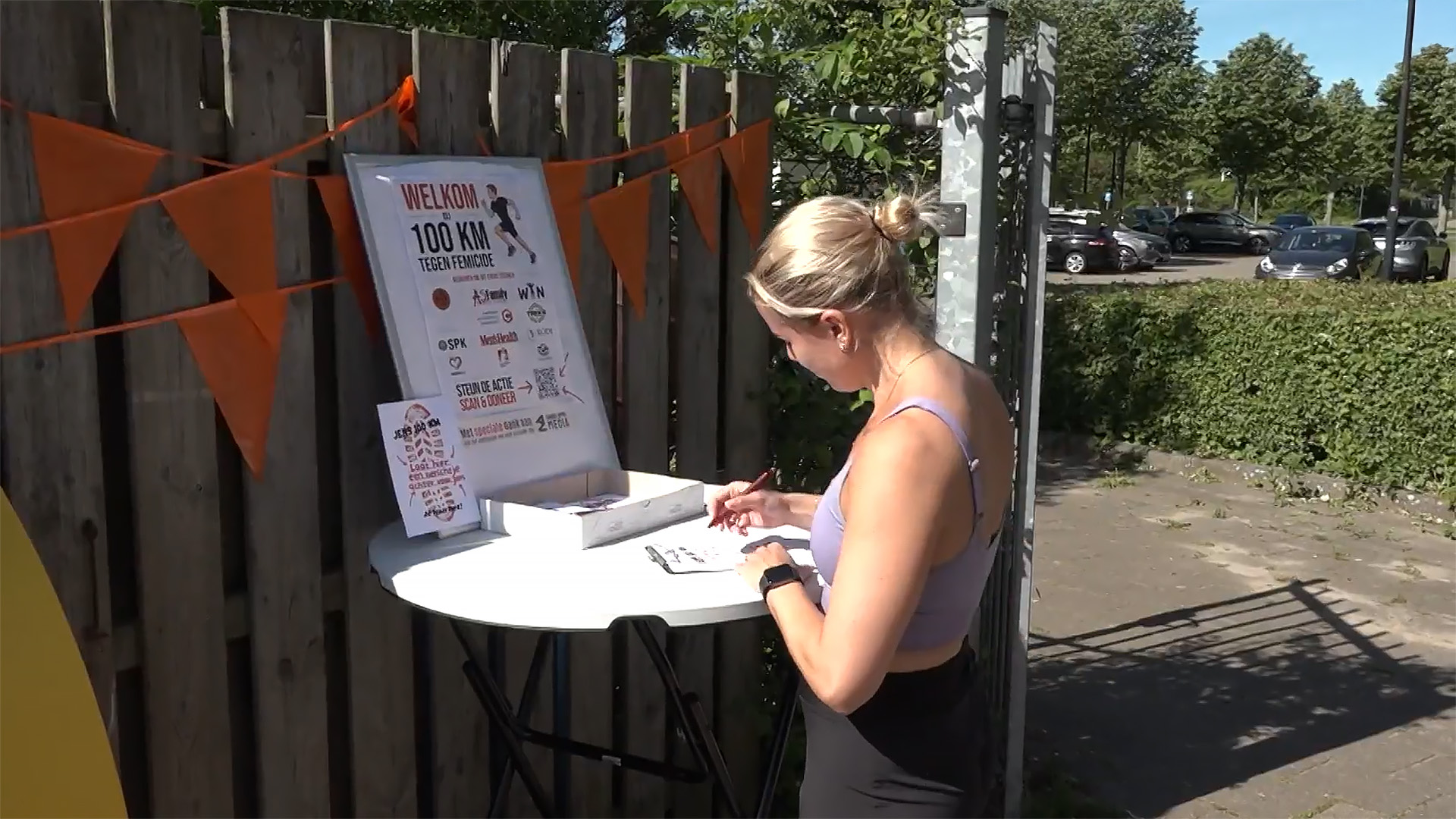 Vrouw schrijft op een formulier op een statafel buiten, bij een bord dat een evenement tegen femicide aankondigt.