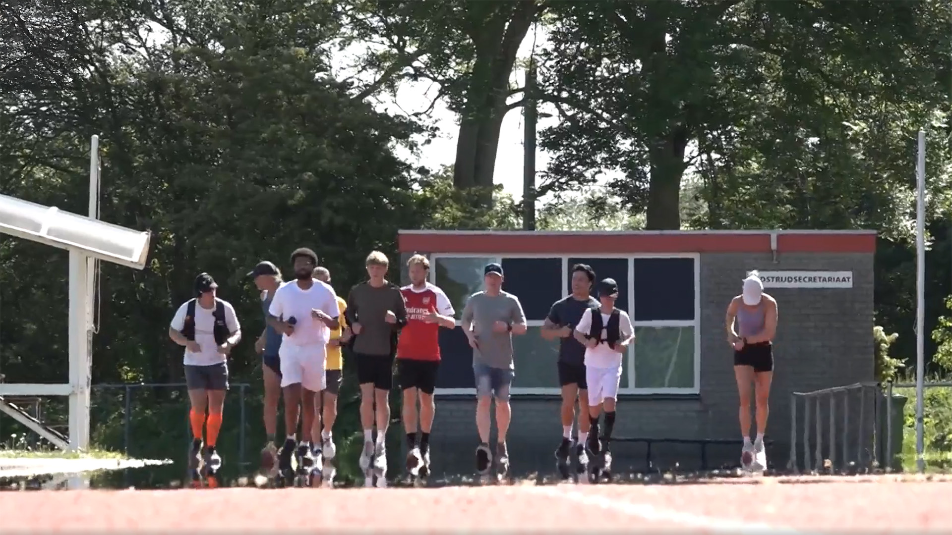 Een groep mensen rent op een atletiekbaan, met bomen en een gebouw op de achtergrond.