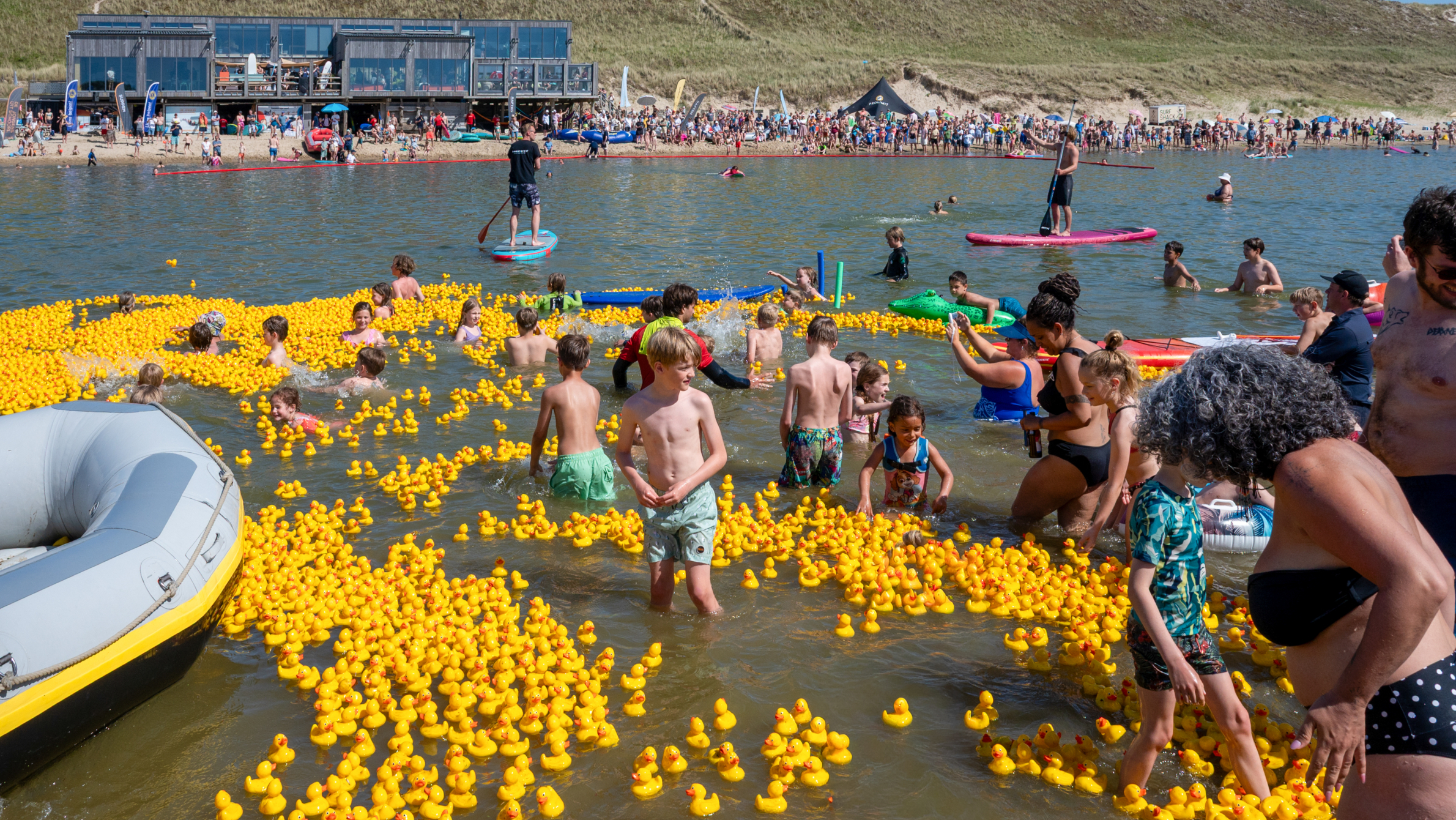 Mensen, veelal kinderen, spelen in het water te midden van een grote hoeveelheid gele rubberen eendjes op een zonnige dag.