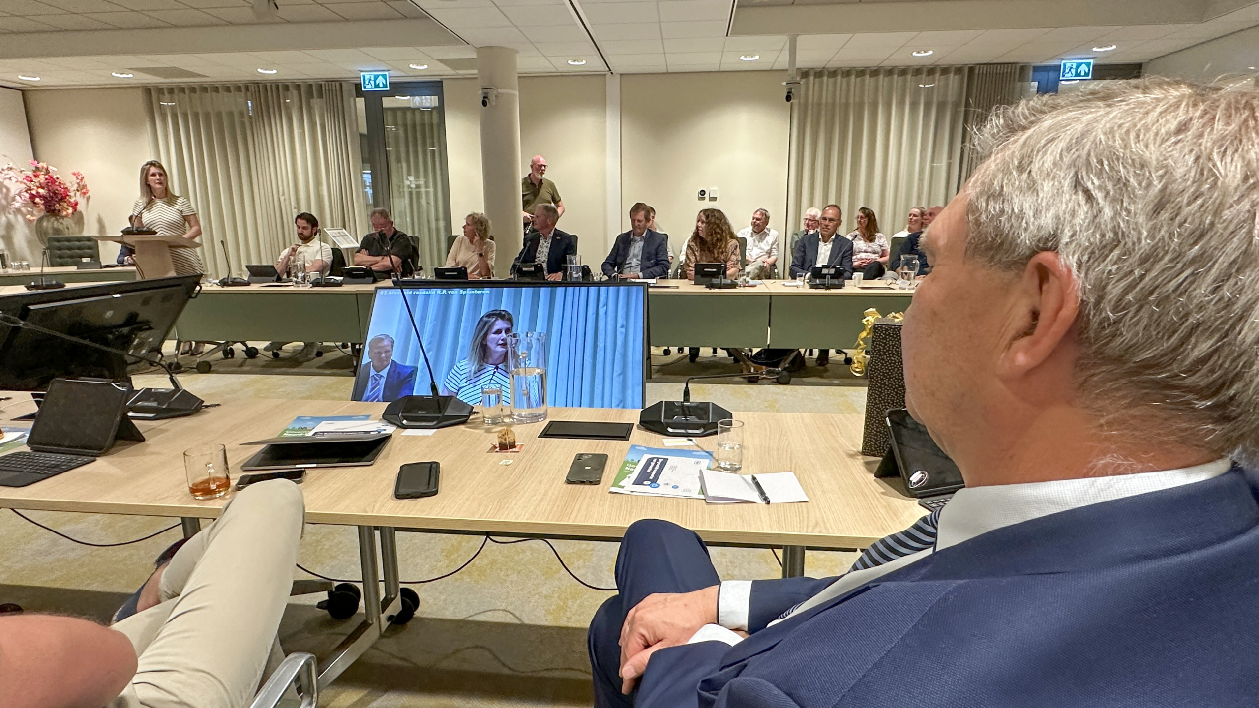 Vergaderzaal met mensen zittend aan tafels, enkele personen spreken of luisteren, en een scherm toont een vrouw die spreekt.