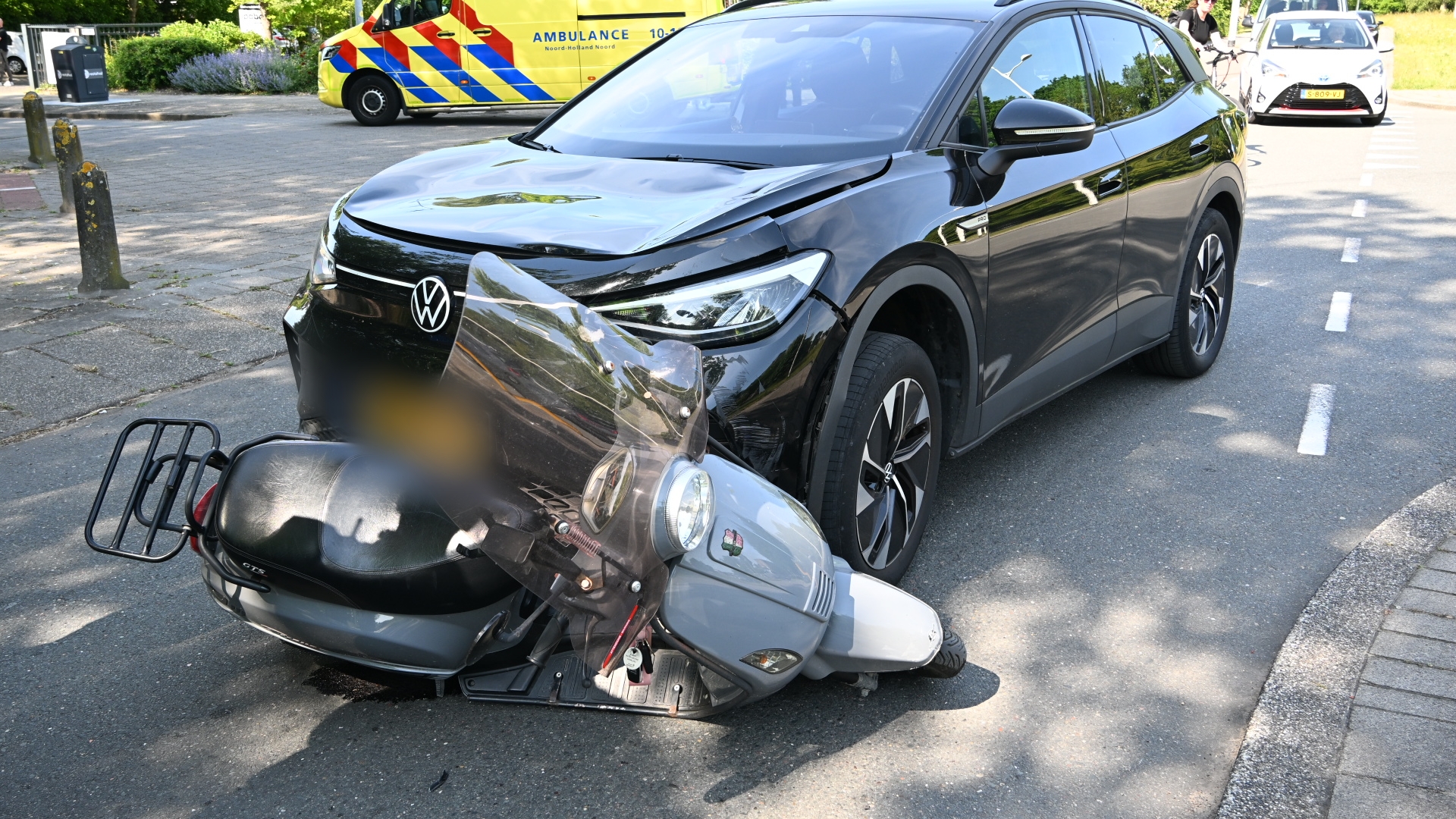 Een zwarte auto is botst met een grijze scooter op een straat; een ambulance staat op de achtergrond.