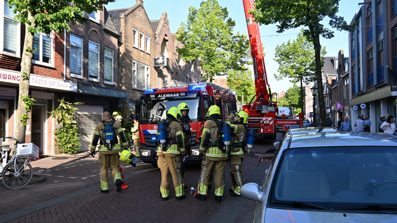 Brandweermannen in gele helmen staan bij een brandweerwagen in een straat met winkels in een rijtje.