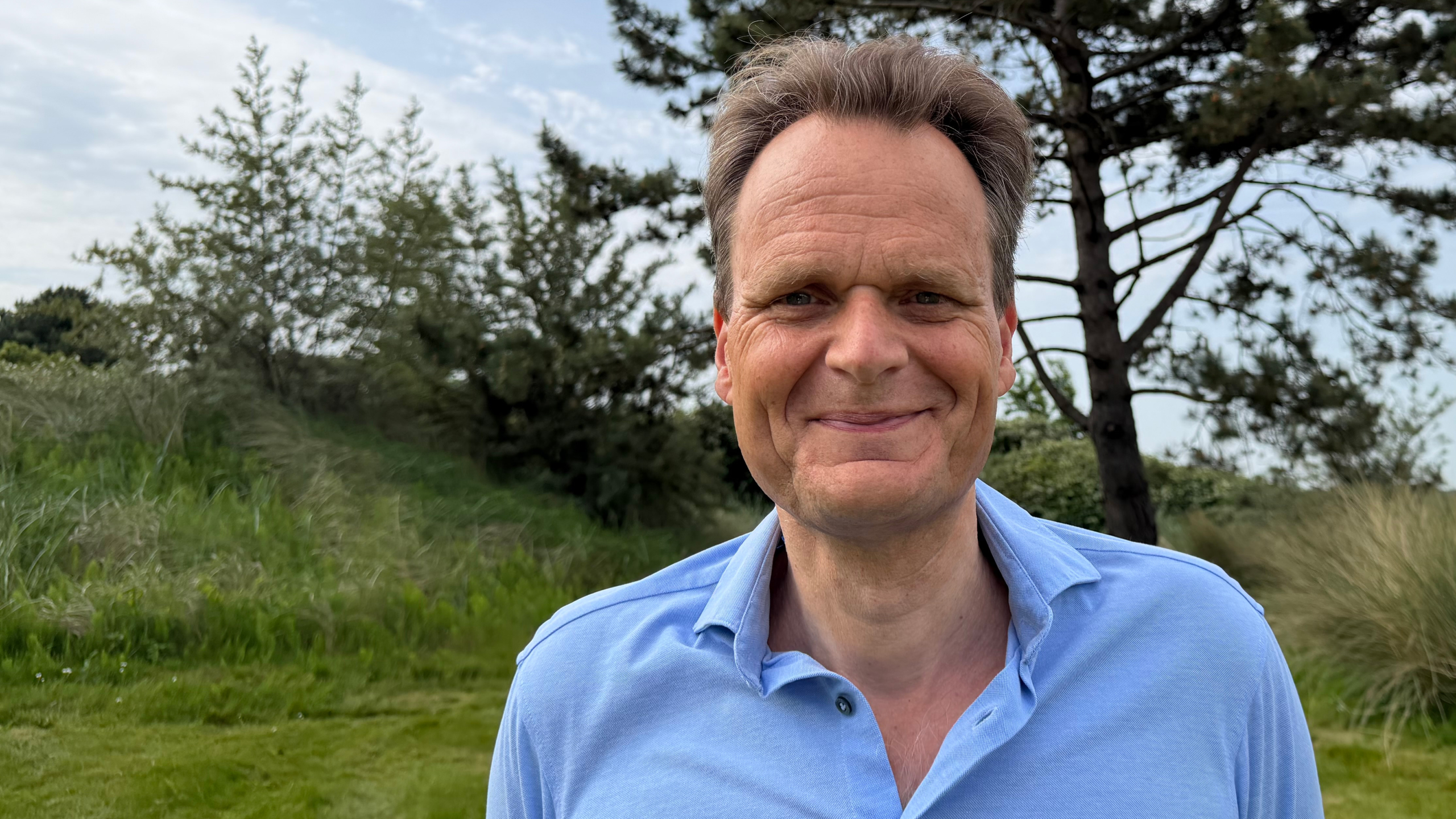 Man in een blauwe polo lacht voor een groene achtergrond met bomen en gras.