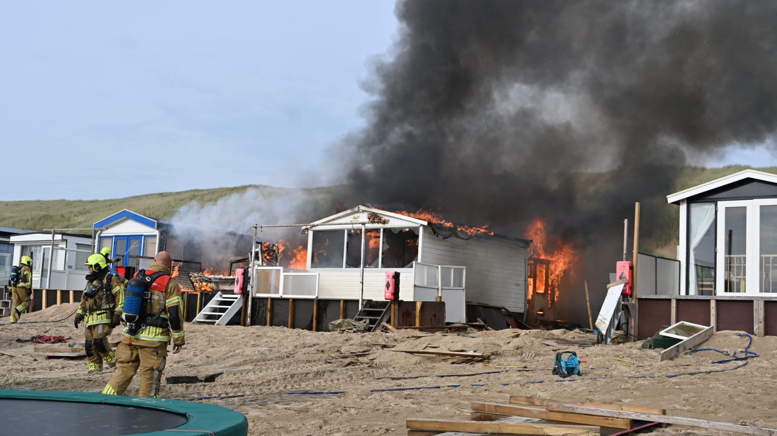 Brandweerlieden bestrijden een uitslaande brand in een strandhuisje, met dikke zwarte rookpluimen stijgend in de lucht.