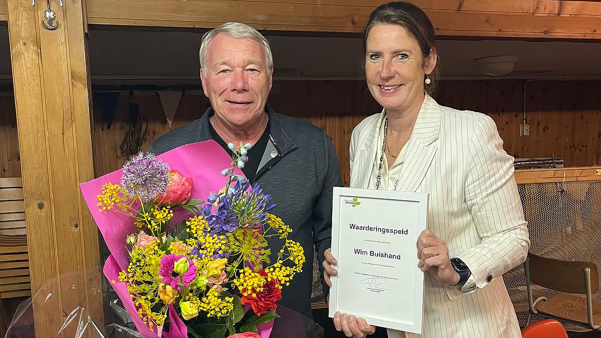 Een man met een boeket bloemen staat naast een vrouw die een oorkonde vasthoudt in een houten kamer.
