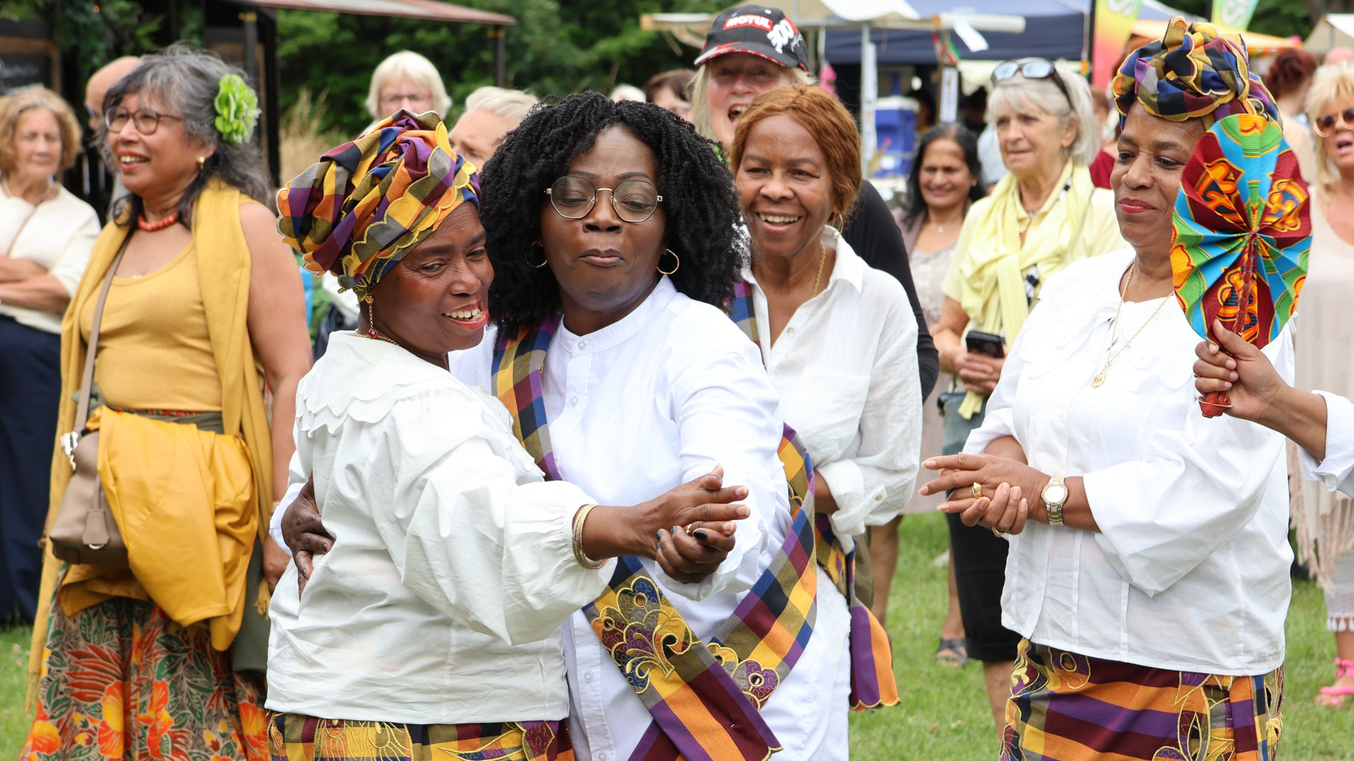 Vrouwen in kleurrijke kleding dansen en lachen op een buitenevenement met toekijkende mensen.