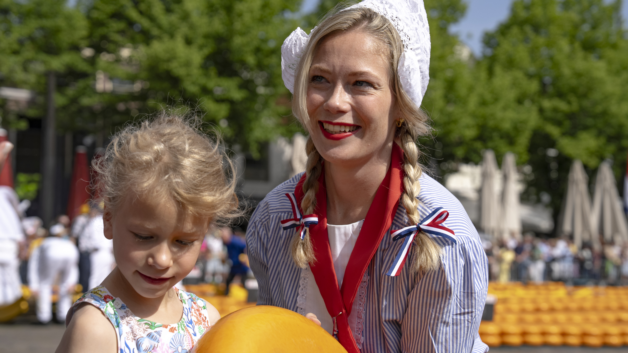 Een vrouw in traditionele Nederlandse kleding glimlacht naast een kind dat een grote kaas vasthoudt op een kaasmarkt.