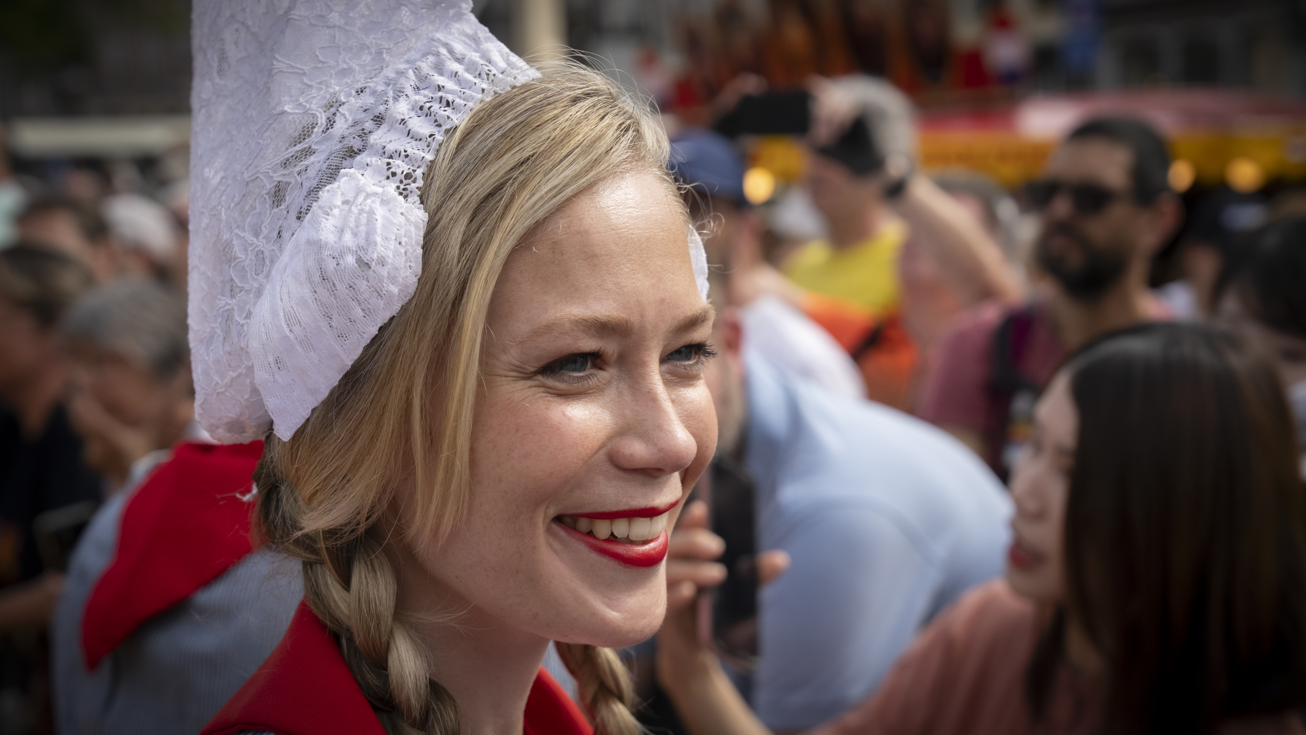Vrouw met vlechten en witte kanten muts lacht in een menigte.