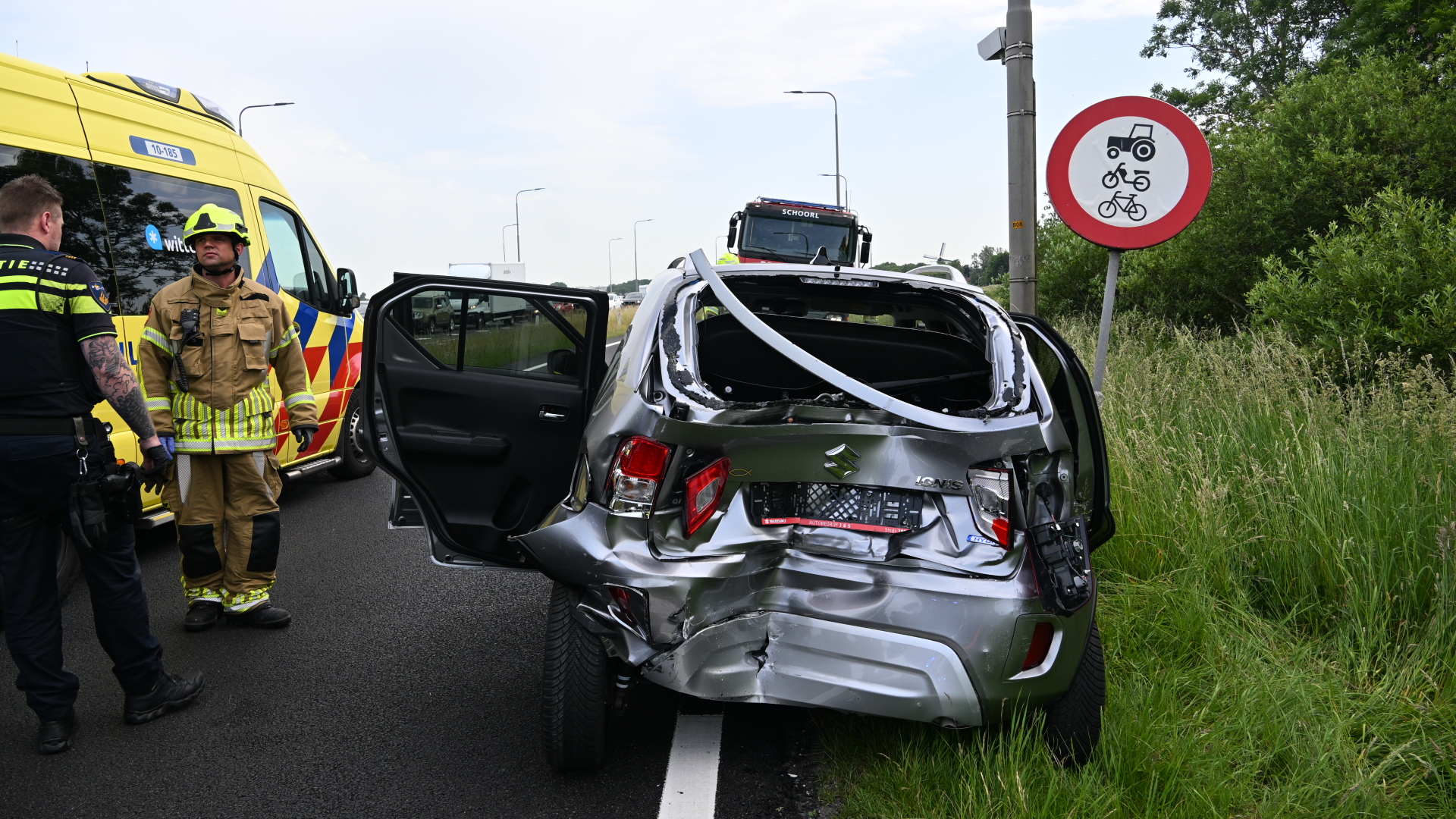 Verfrommeld voertuig betrokken bij ongeval langs de weg, met een brandweerman en een hulpverlener naast een ambulance.
