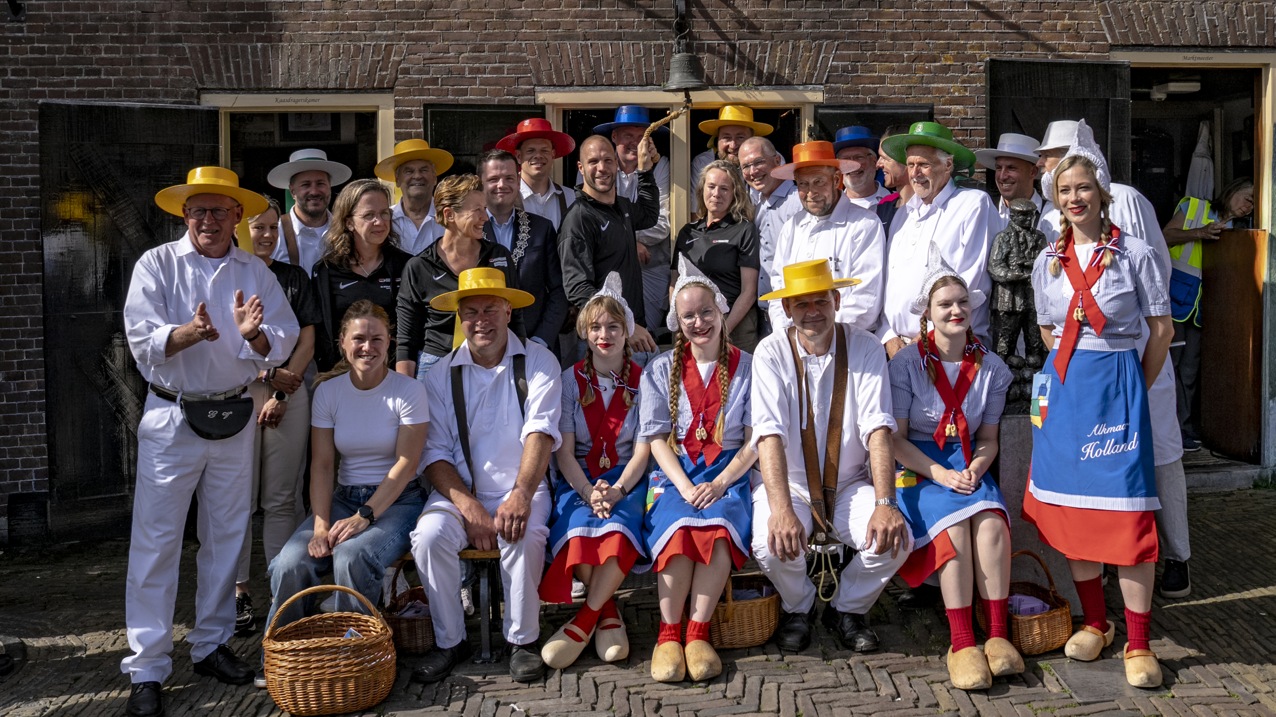 Groep mensen in traditionele Nederlandse kleding voor een bakstenen gebouw, sommigen dragen kleurrijke hoeden en klompen.