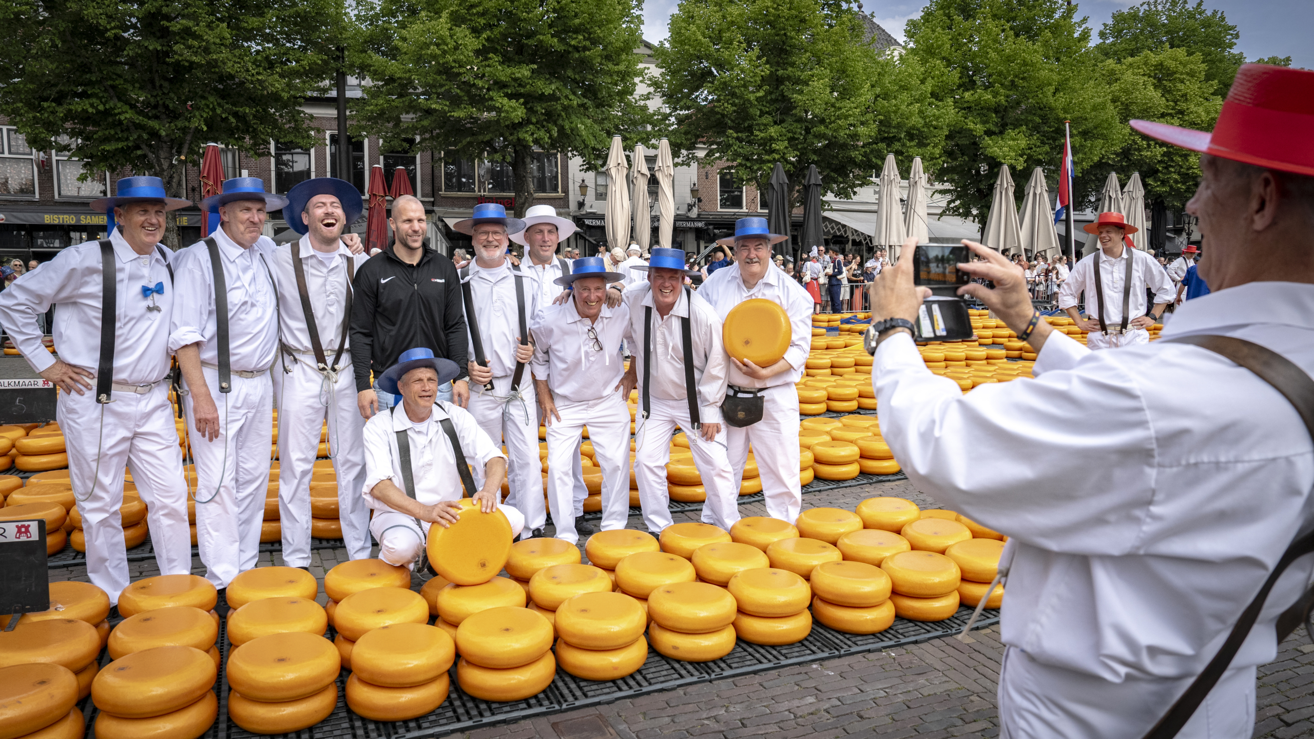 Mensen in witte kleding poseren met grote gele kazen op een markt, terwijl een man een foto van hen maakt.
