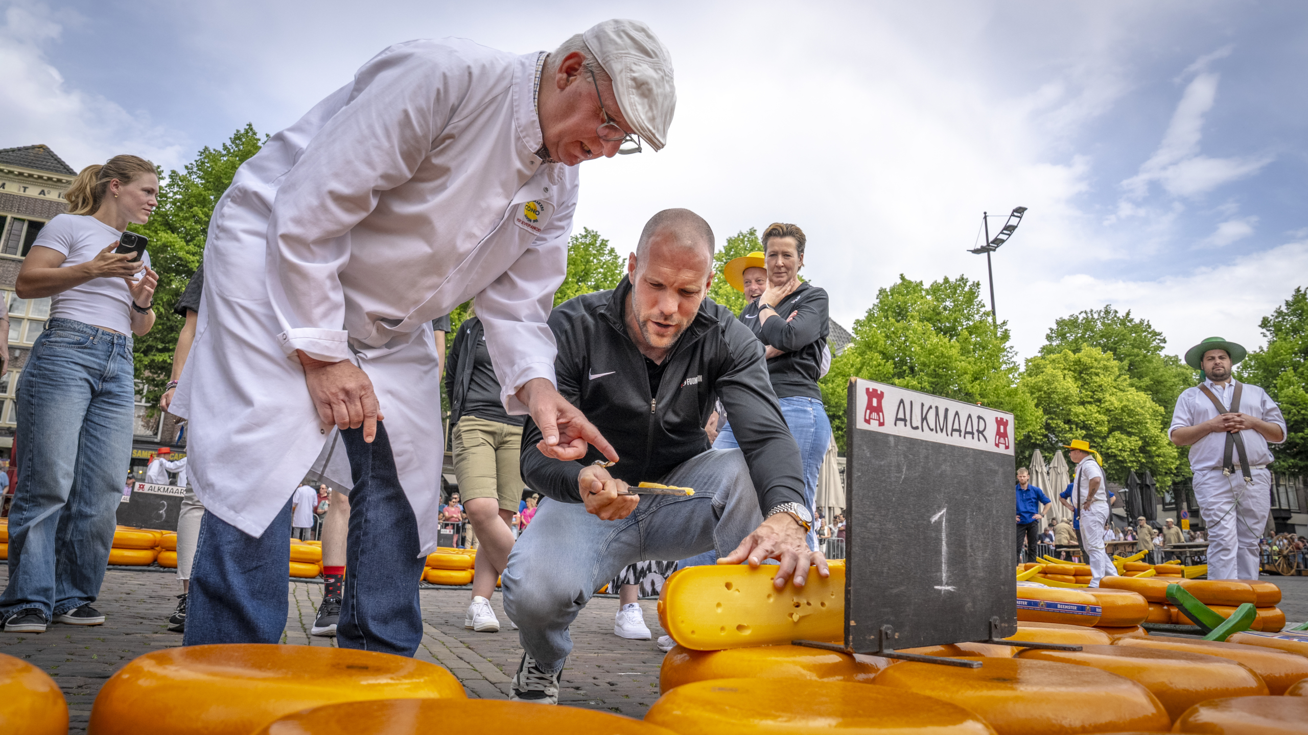 Mensen keuren kazen op de kaasmarkt in Alkmaar, met een man die een kaasboring onderzoekt onder toezicht.