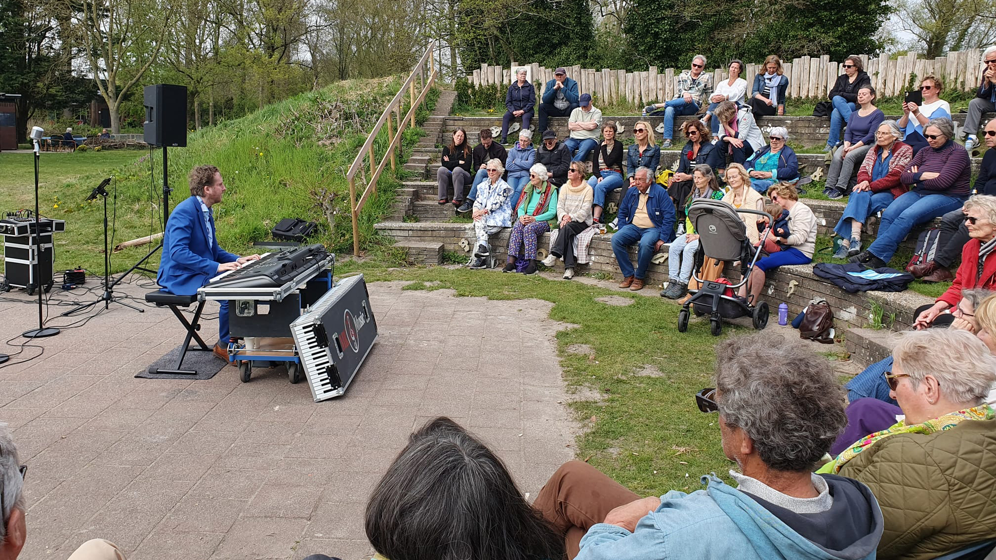 Een pianist in een blauw pak speelt voor een zittend publiek in een openluchttheater met groene omgeving.