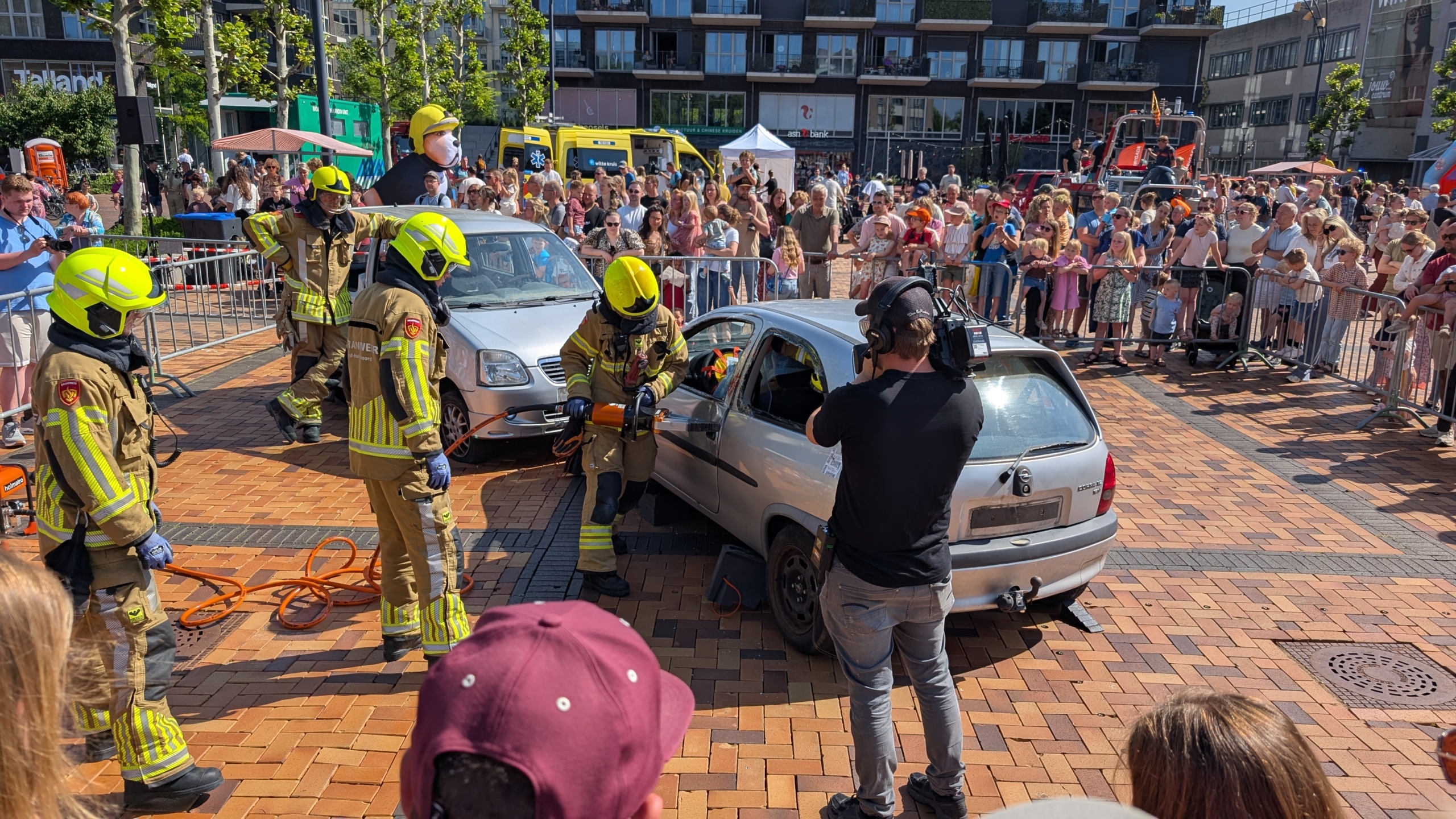 Brandweerlieden demonstreren reddingswerkzaamheden bij een auto-ongeluk voor een publiek op een zonnig plein.