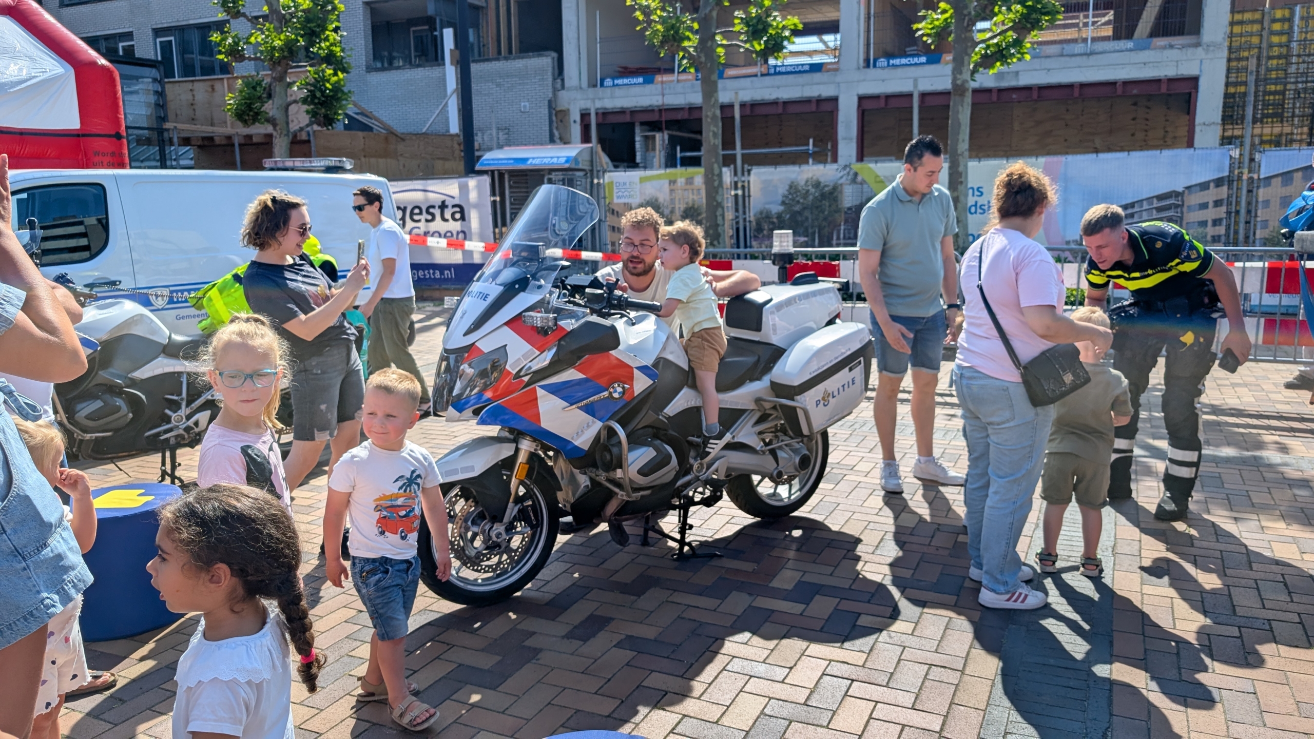 Kinderen bewonderen een politie-motorfiets op een plein, terwijl een agent met familieleden praat.