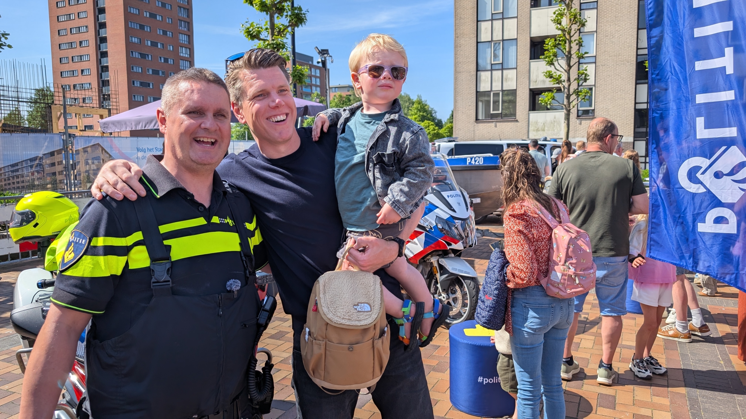 Een politieagent poseert glimlachend met een man die een jong kind in een drukke stedelijke omgeving vasthoudt.