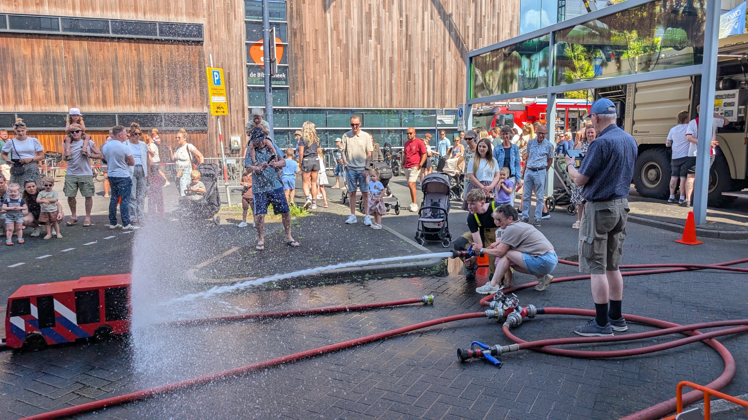 Kinderen spuiten water met een brandslang, omringd door toeschouwers op een zonnige dag bij de brandweer.