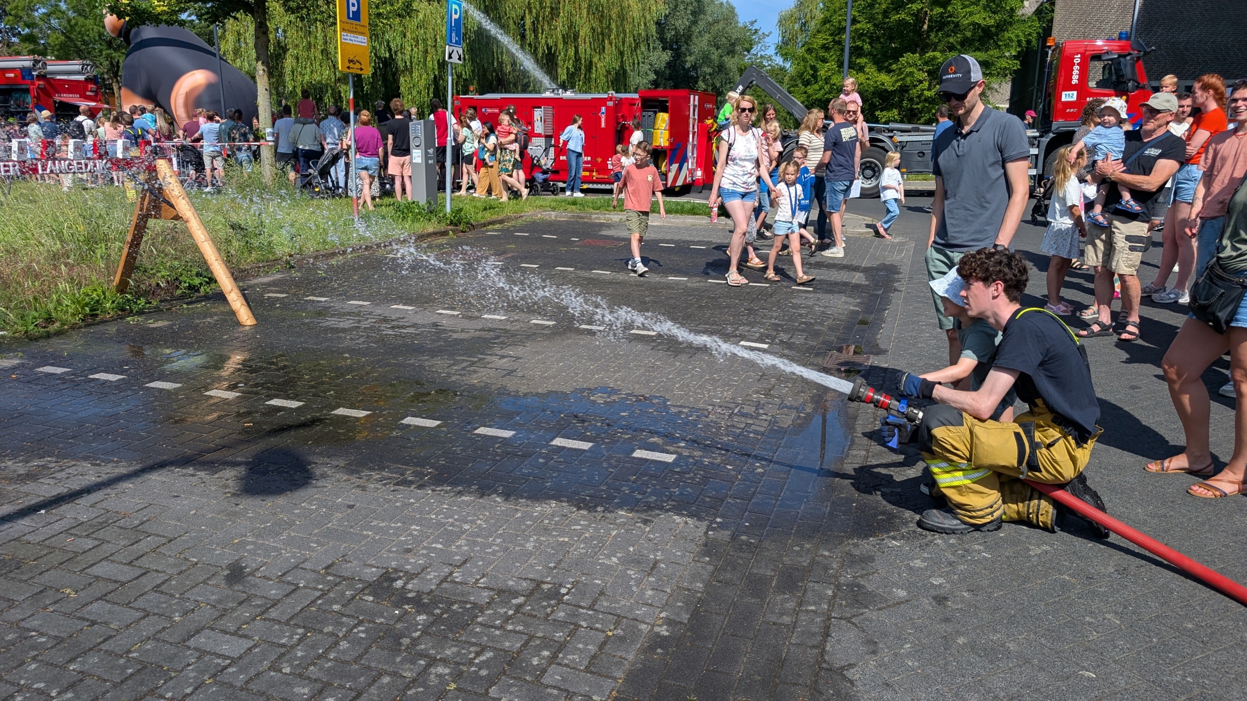 Brandweerman spuit water met een brandslang, omringd door toekijkende mensen bij een open dag met brandweerwagens.