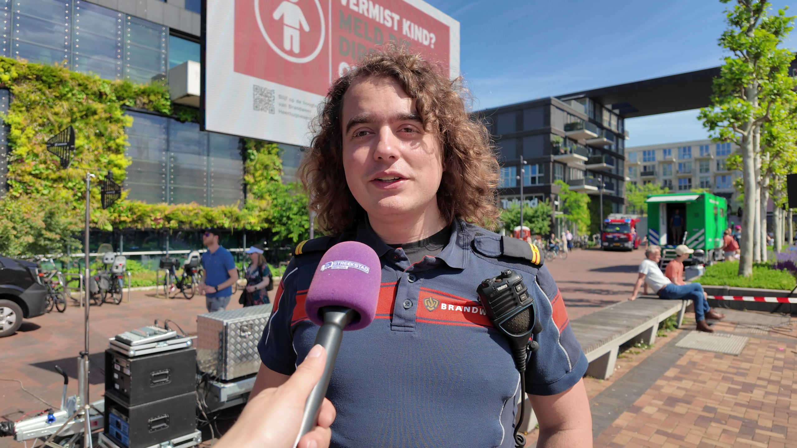 Een brandweerman met krullend haar wordt geïnterviewd op een zonnige dag in een stedelijk gebied met omstanders en apparatuur.