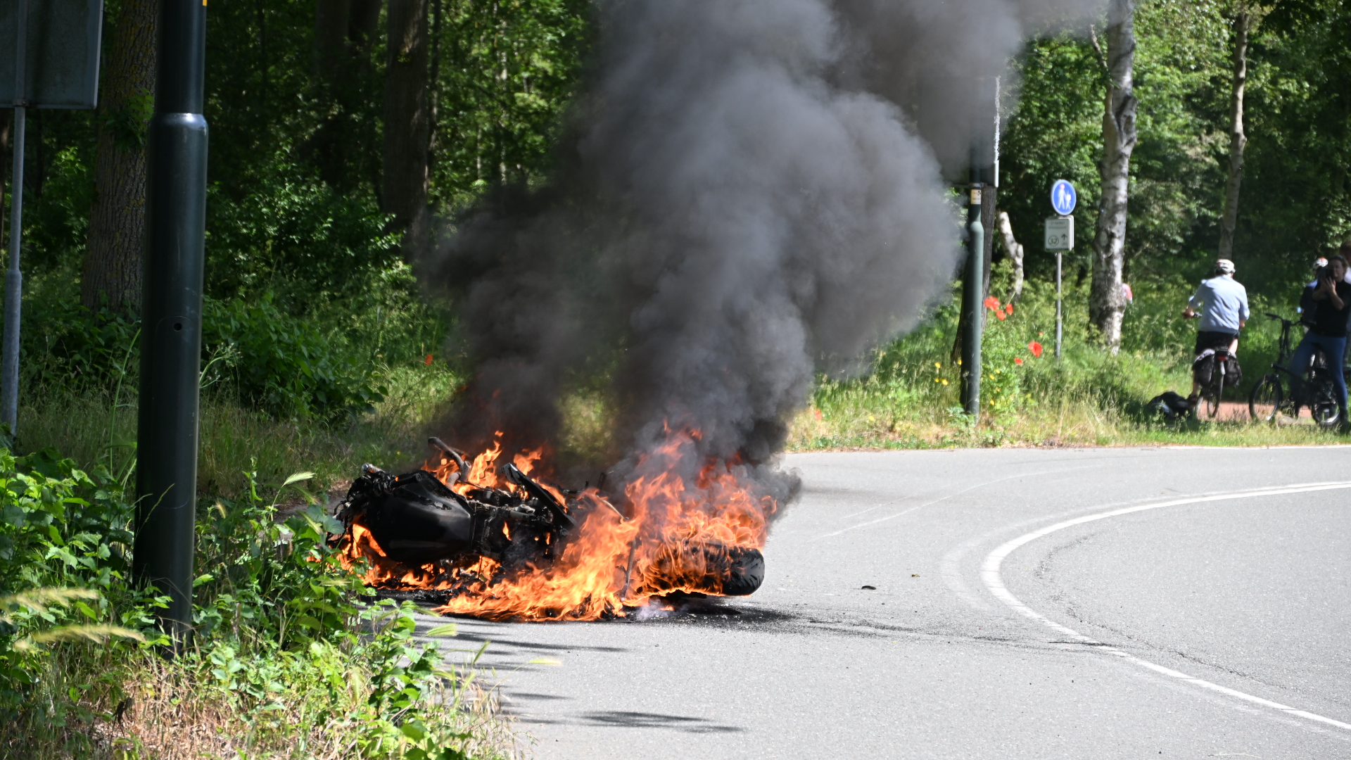 Brandende motorfiets op een weg met dikke zwarte rook, omgeven door groen; enkele toeschouwers op de achtergrond.