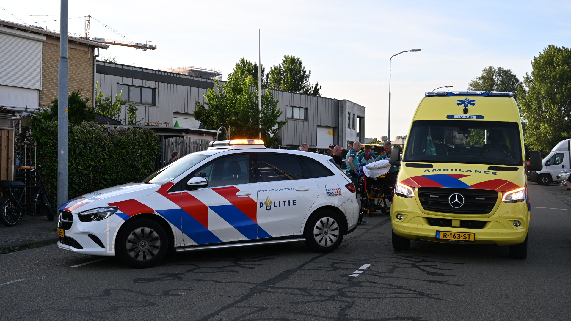 Politie- en ambulancevoertuig staan geparkeerd op straat naast mensen met een brancard, waarschijnlijk bij een noodsituatie.