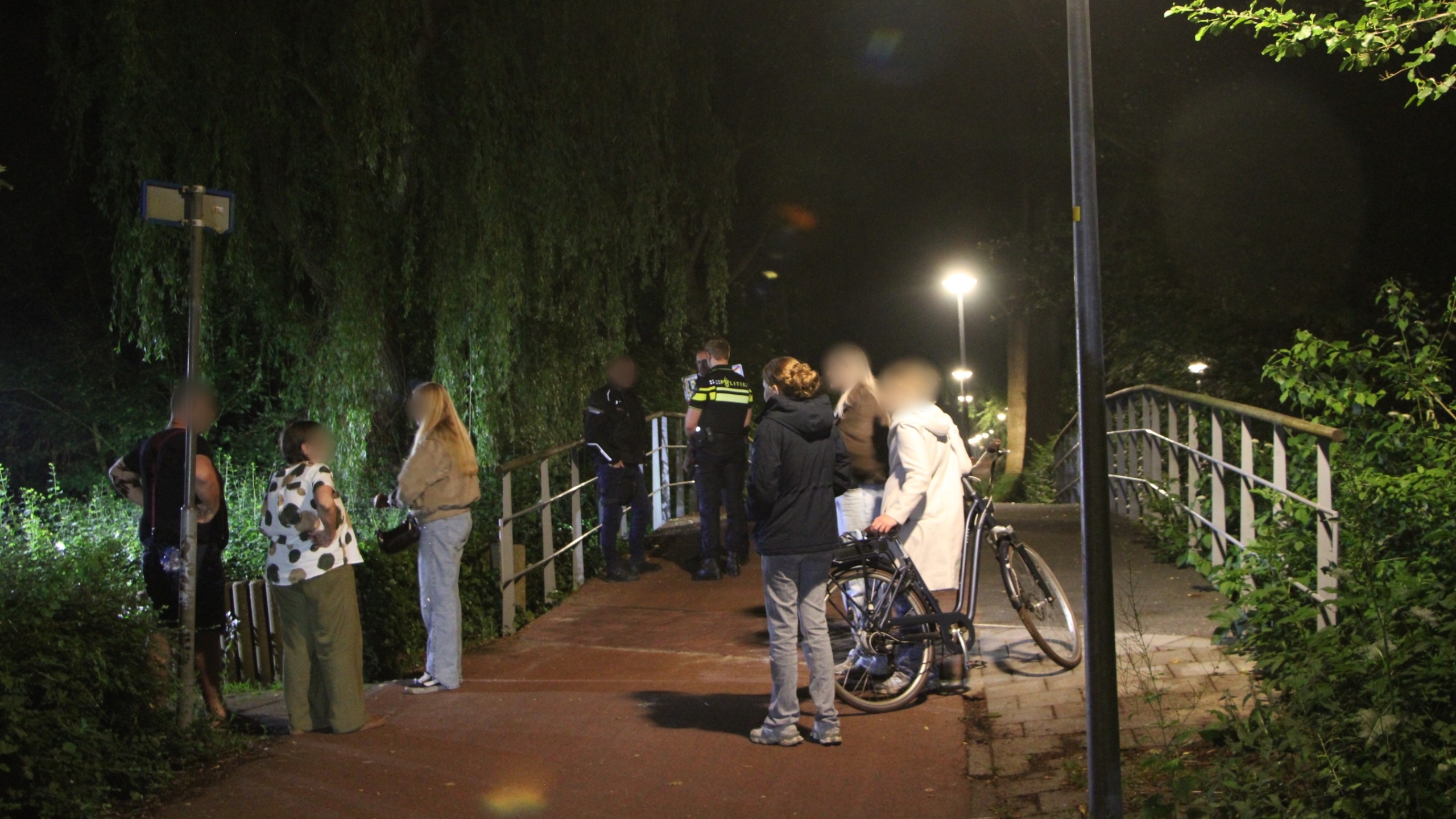 Groep mensen, inclusief een politieagent, staan 's nachts op een verlicht pad naast een brug en fiets.