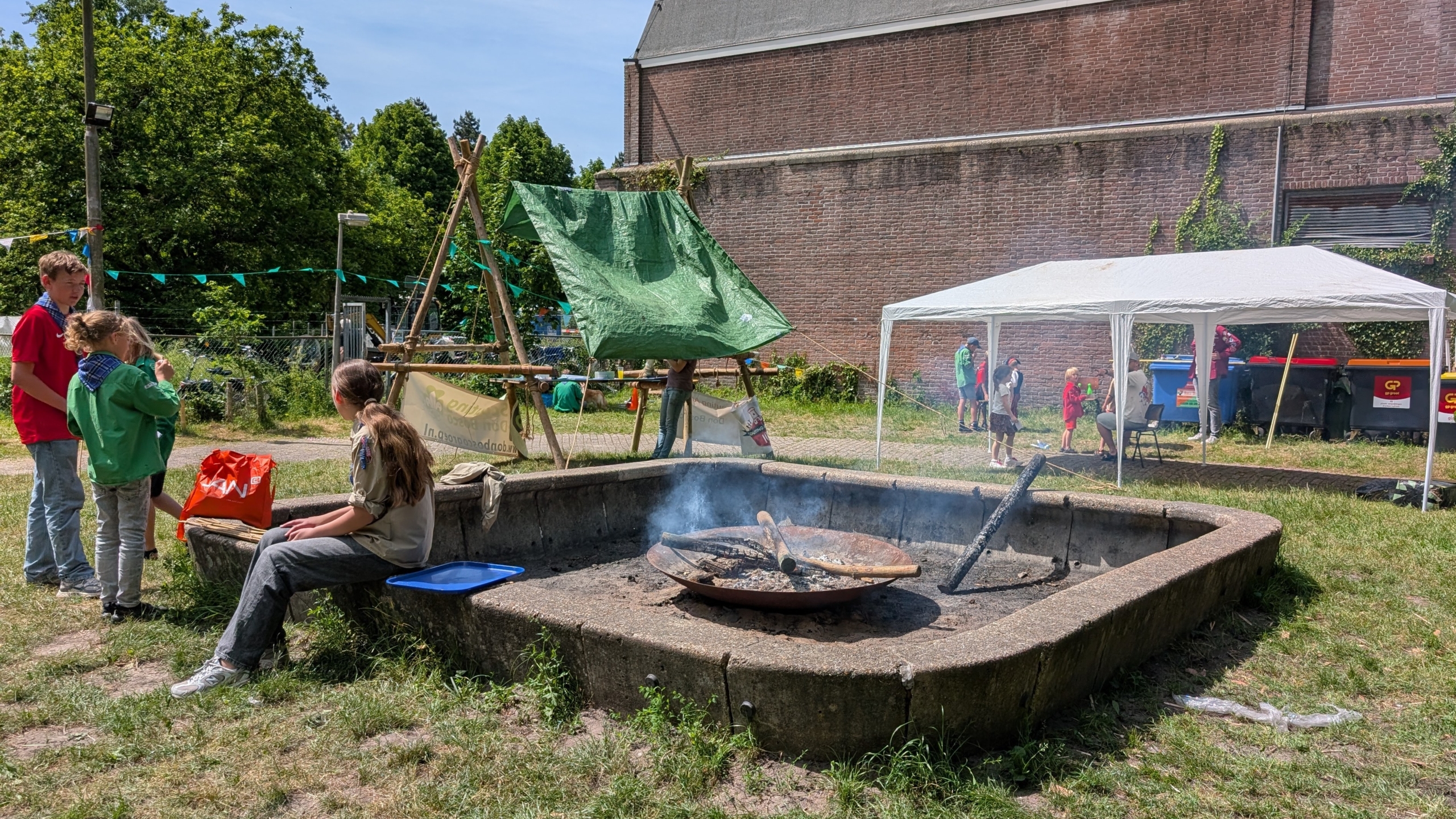 Een groep mensen is buiten bij een vuurplaats met een smeulend vuur; er zijn ook een wit tentje en een groene afdekzeil zichtbaar.