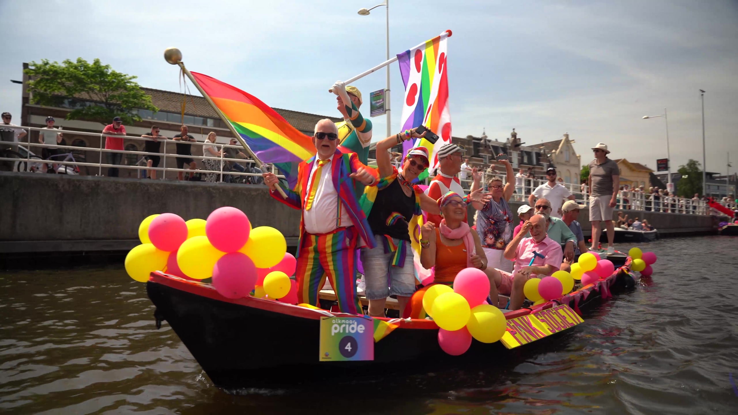 Mensen in kleurrijke kleding vieren op een met ballonnen versierde boot tijdens Alkmaar Pride, met regenboogvlaggen.