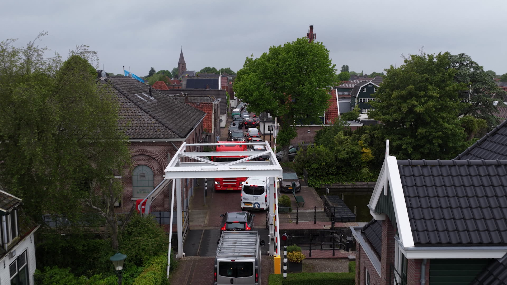 Een filesituatie op een smalle straat omringd door huizen, een wit ophaalbruggetje, en een groene boom op de achtergrond.