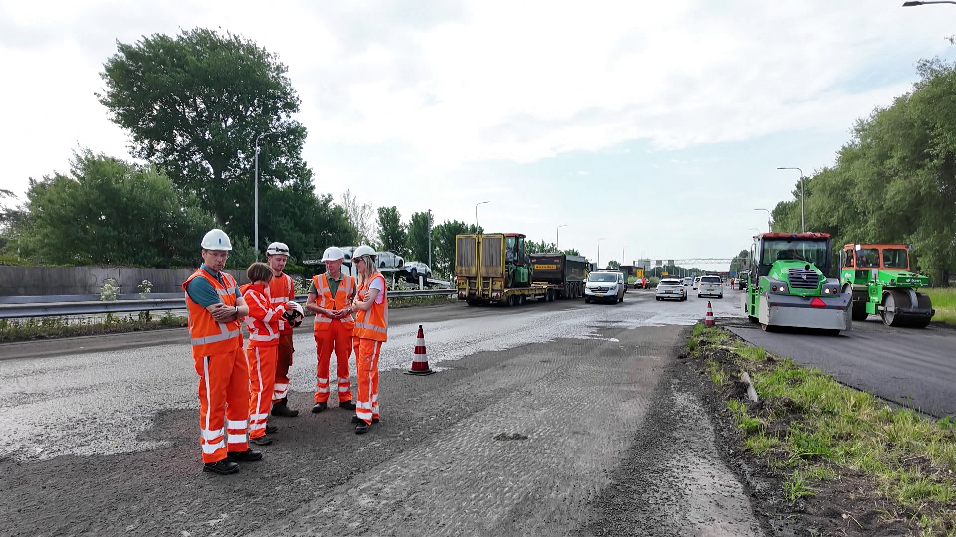 Werknemers in oranje veiligheidskleding en helmen staan op een weg bij een wegenbouwproject, omgeven door machines en auto's.