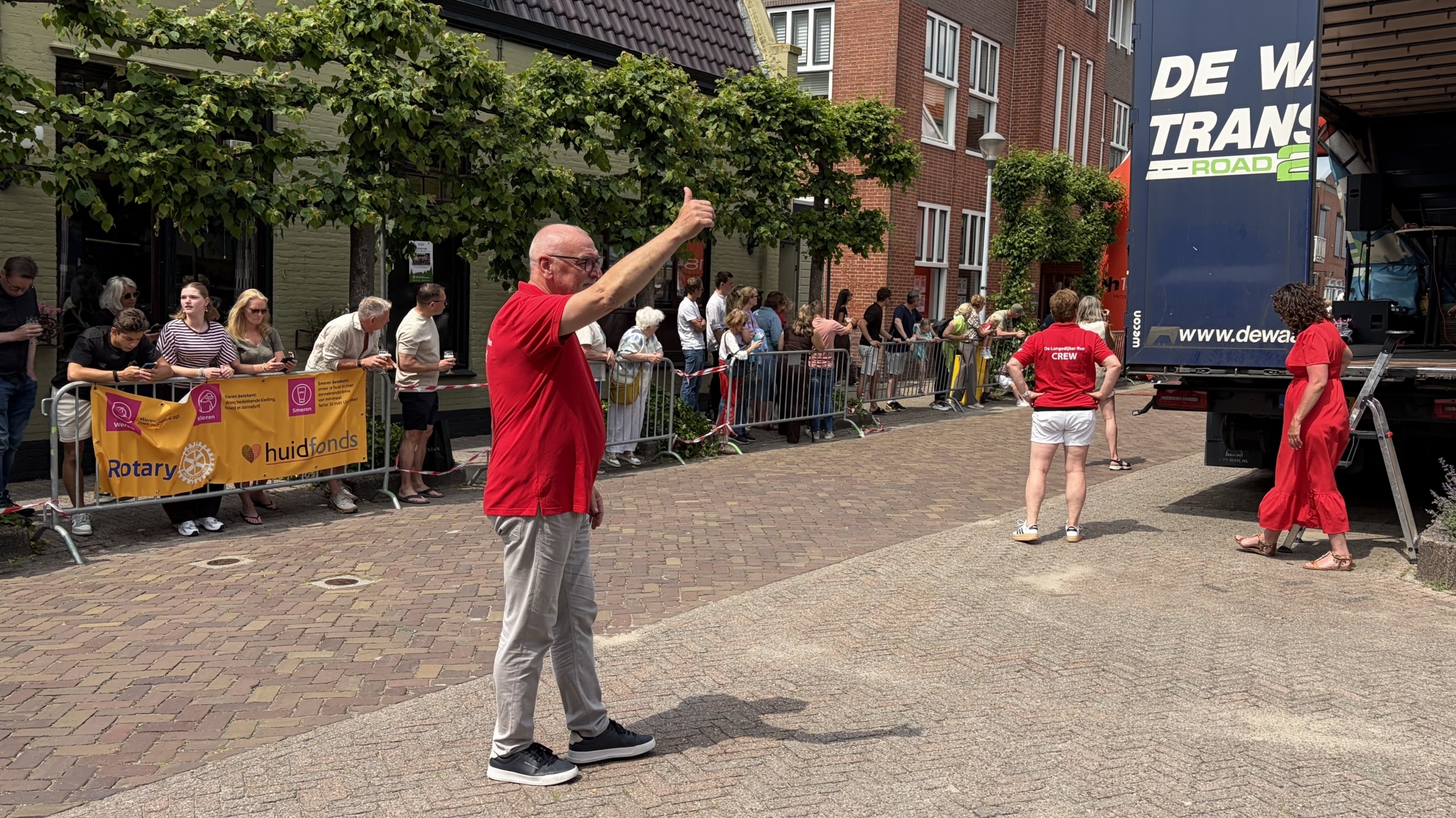 Een man in een rood shirt steekt zijn duim omhoog naast een vrachtwagen tijdens een evenement met toeschouwers en vrijwilligers in de buurt.