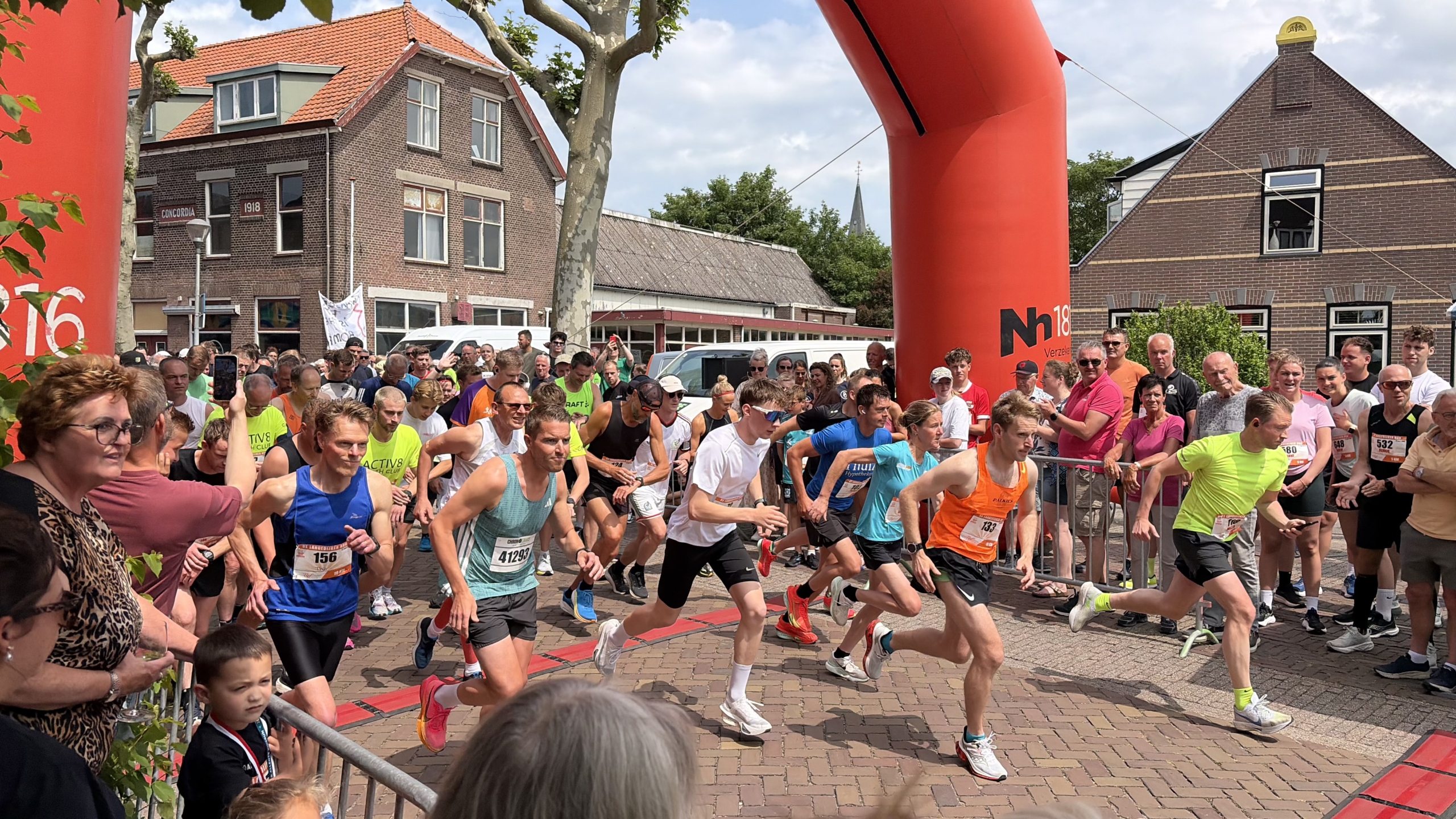 Hardlopers starten een race onder een grote rode boog, omringd door toeschouwers en gebouwen.